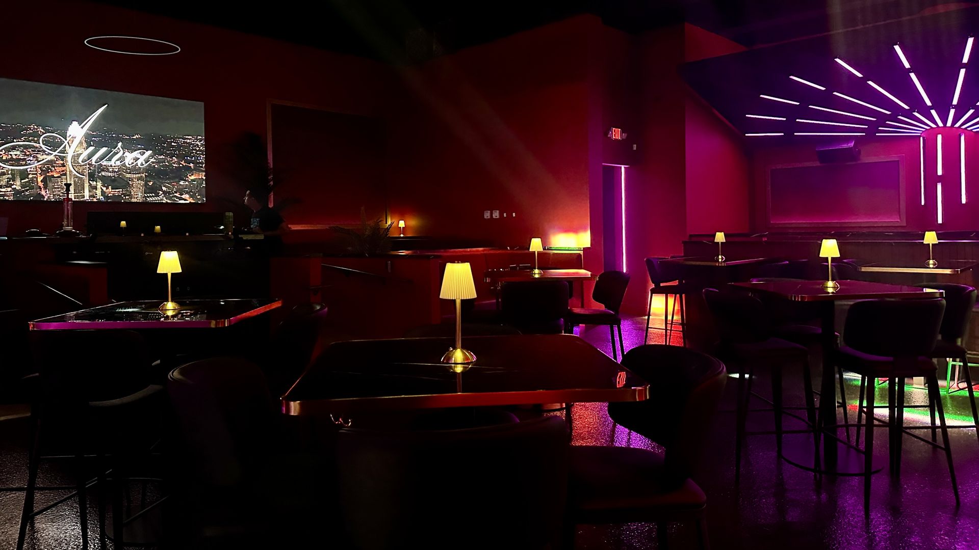 Dimly lit lounge with dark tables, black chairs, and yellow lamps. Pink and purple neon lights on walls and ceiling, with a screen displaying the word "Aura" in cursive.