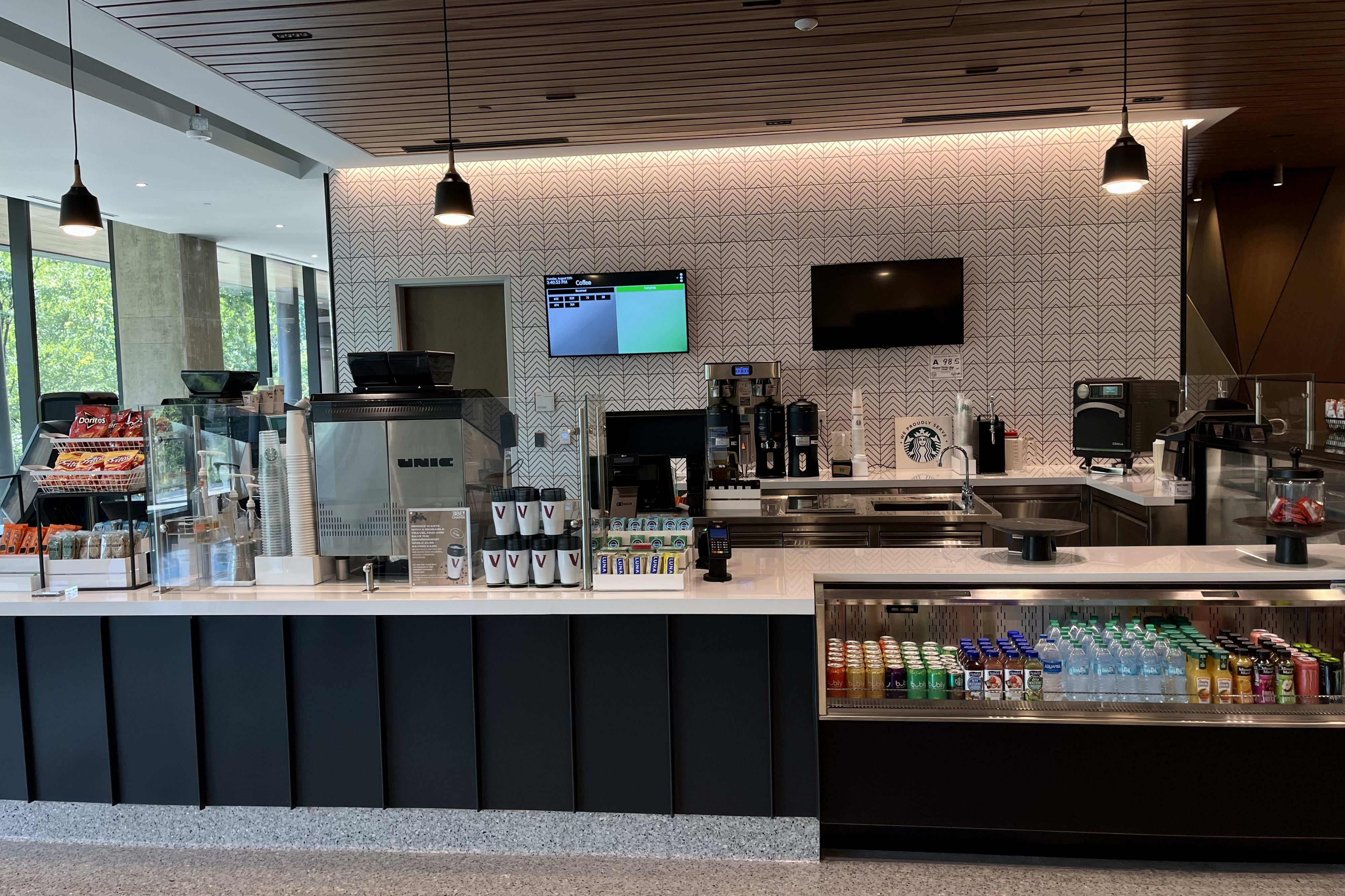 Modern coffee shop counter with snack racks, coffee cups, beverage fridge with water and soda, coffee machines, and two digital screens on a patterned wall under hanging black lights.