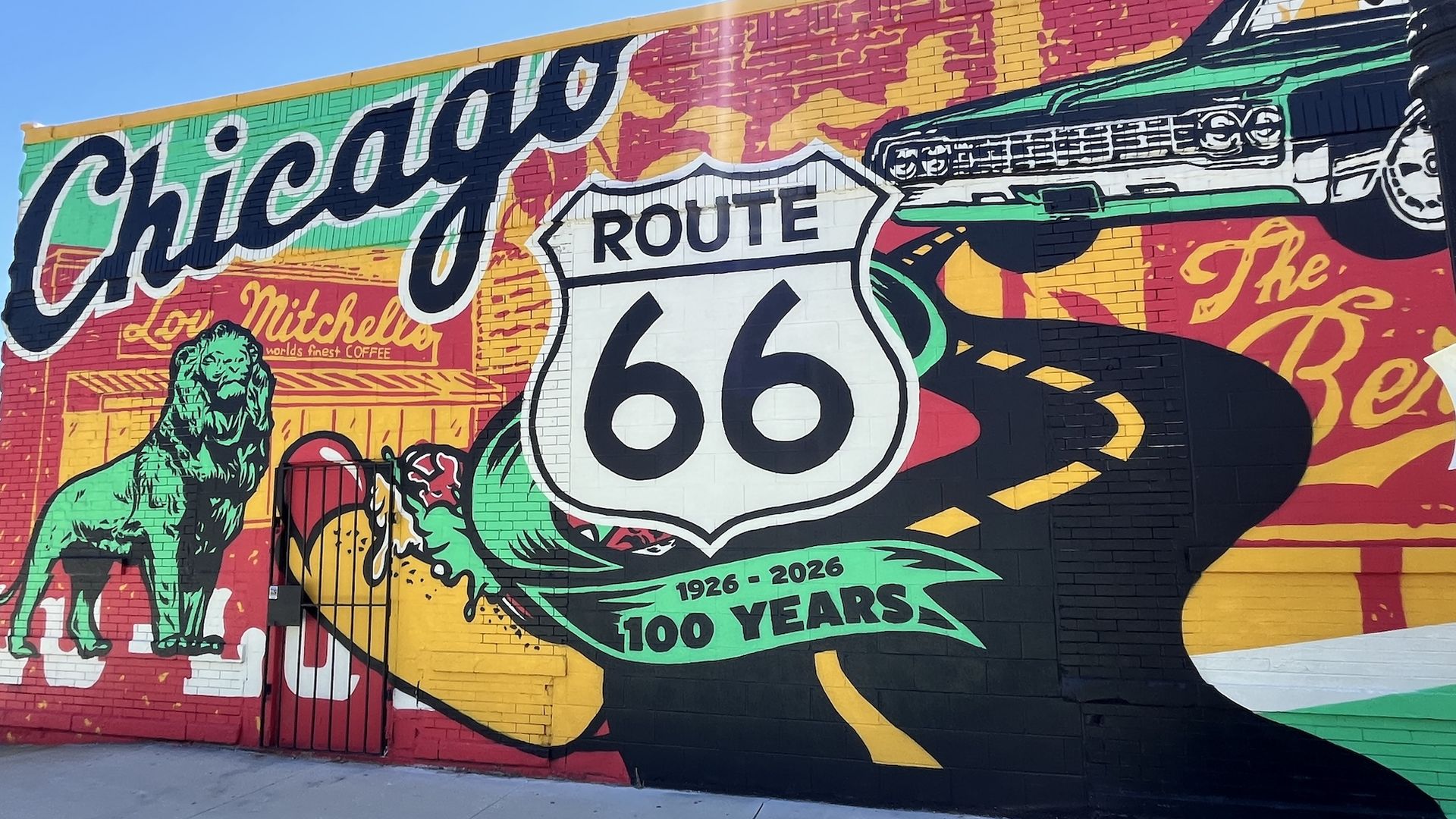 Vibrant mural on a brick wall celebrating Route 66: Chicago in large cursive, Route 66 shield, a vintage car, and the years 1926-2026 in green and yellow, against a blue sky.