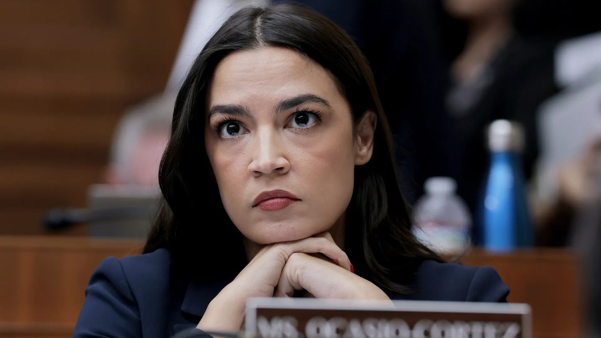 Photo of Rep. Alexandria Ocasio-Cortez at a Congressional hearing with her chin in her folded hands