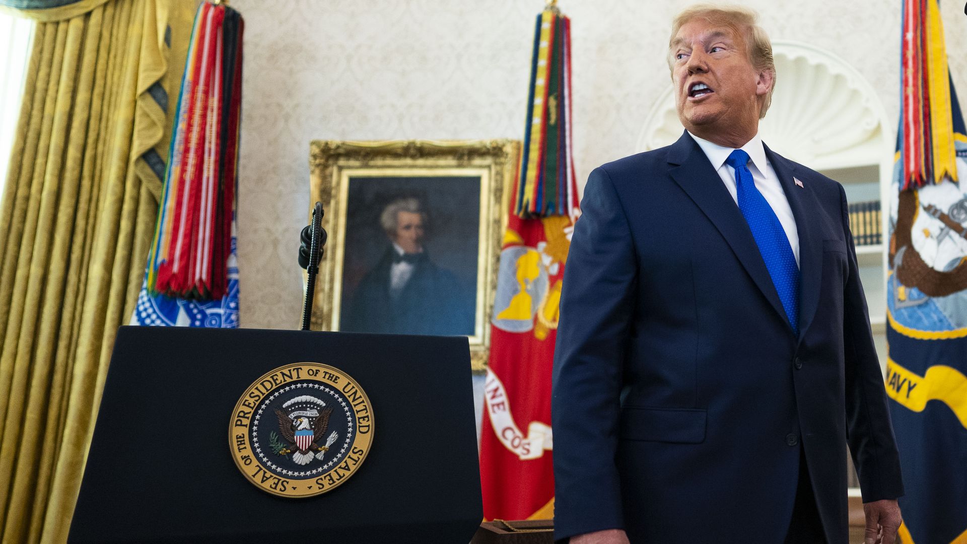Trump wears a suit while standing next to a podium with the presidential seal on it