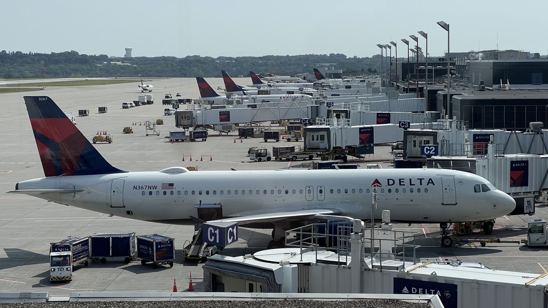 Wide view of a sunny, blue-sky airport gate area: multiple Delta jets docked at gates with jet bridges, ground support vehicles, a distant control tower, and a Delta tail in the foreground.
