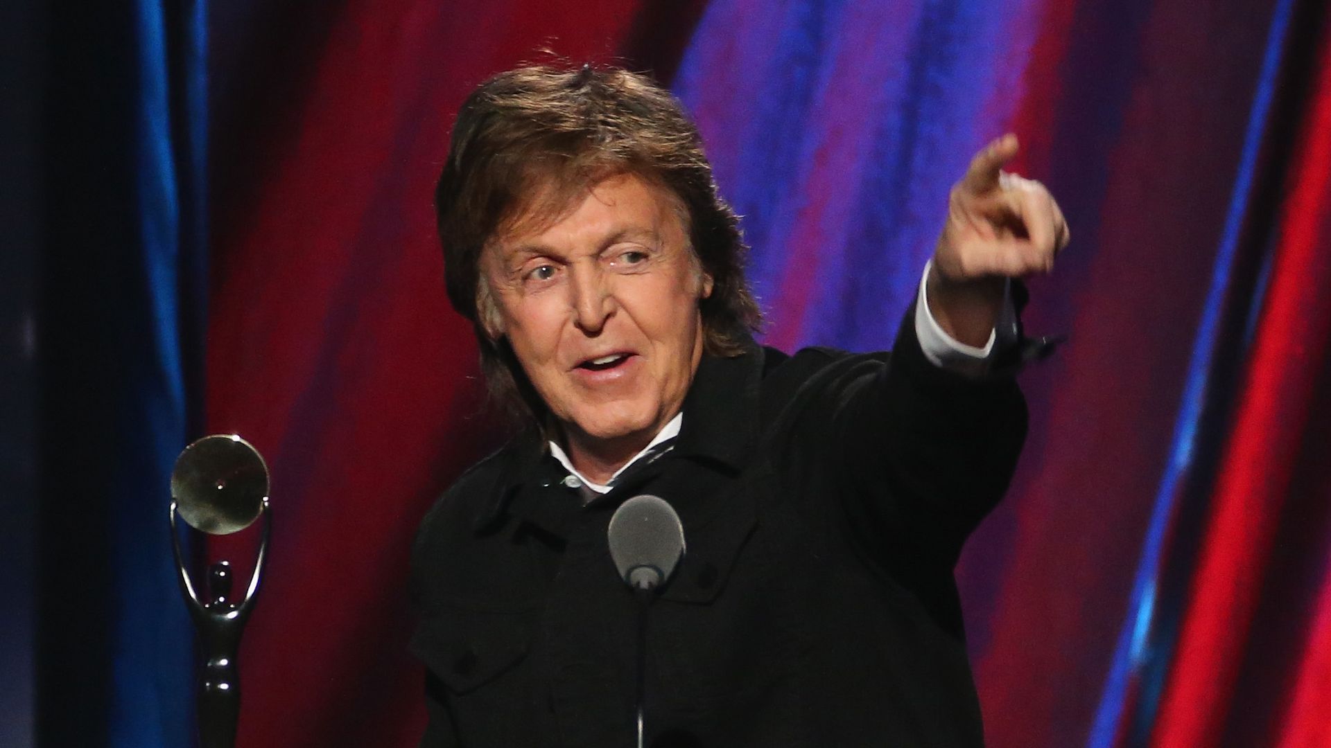 Man with brown hair wearing a black jacket speaks at a microphone and points forward, with a trophy on a podium and red and blue curtains in the background.