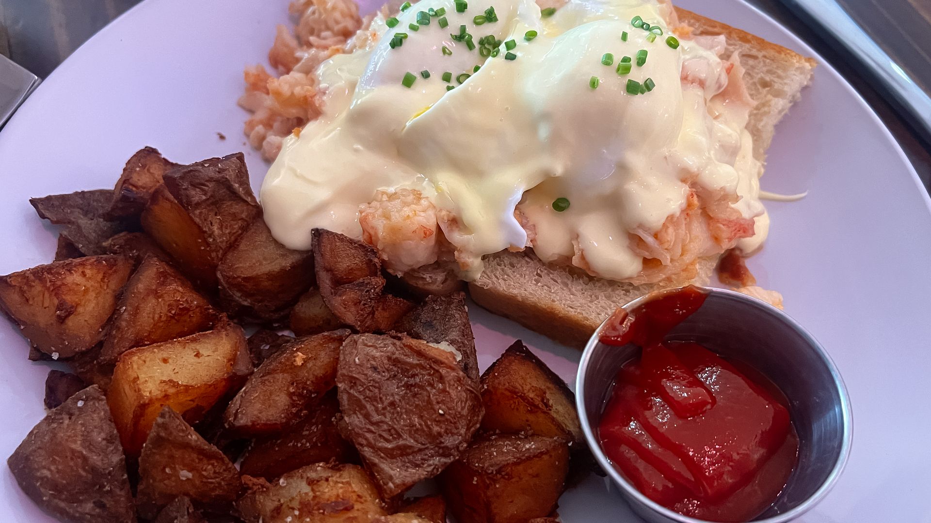 A plate with a lobster Benedict, potatoes and ketchup.