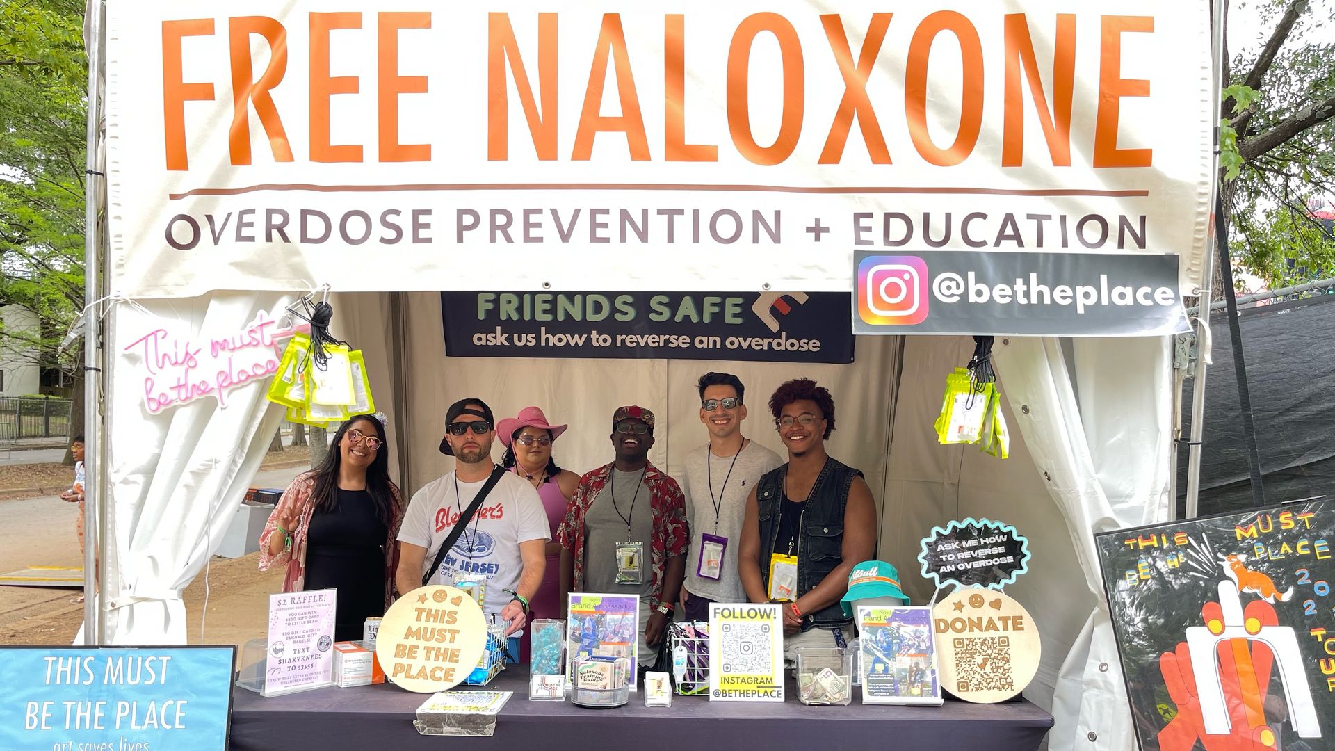 A white banner that reads "Free Naloxone" over a group of people inside a tent, surrounded by books and posters.