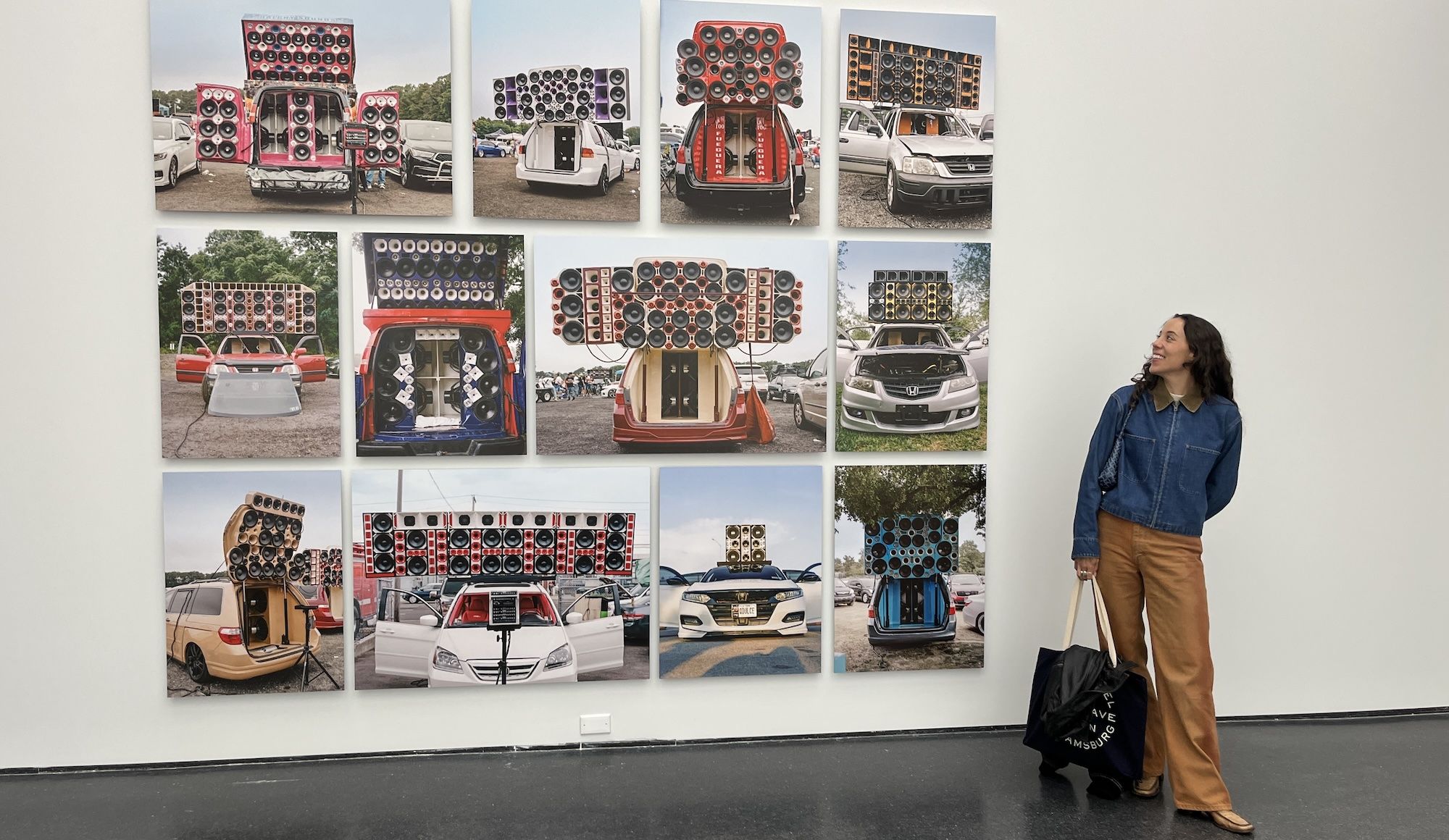 White gallery wall displaying a grid of vehicle photographs with giant speaker setups on car trunks; a woman in a denim jacket and tan pants stands at right, admiring the art.