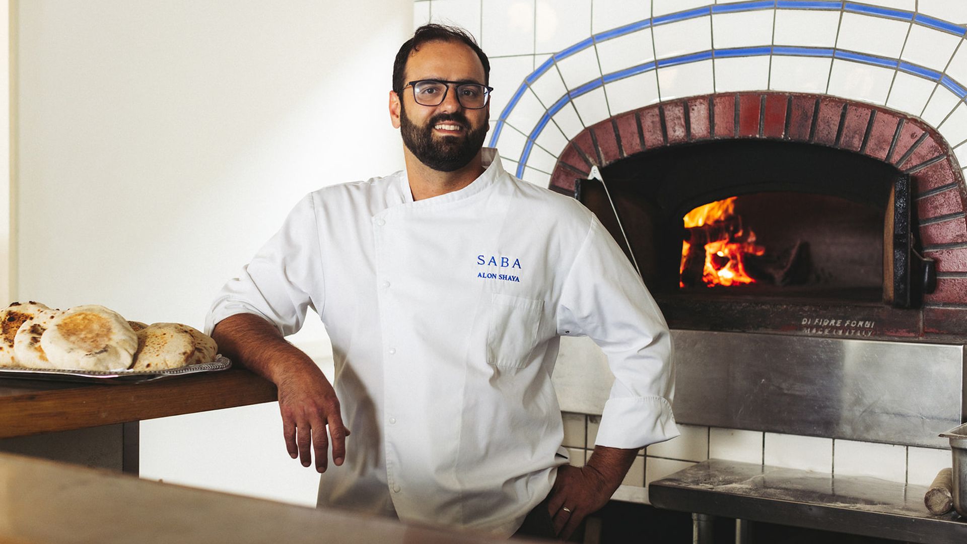 Alon Shaya poses for a photo in front of a wood-burning oven. He leans an elbow on a countertop with pita bread.