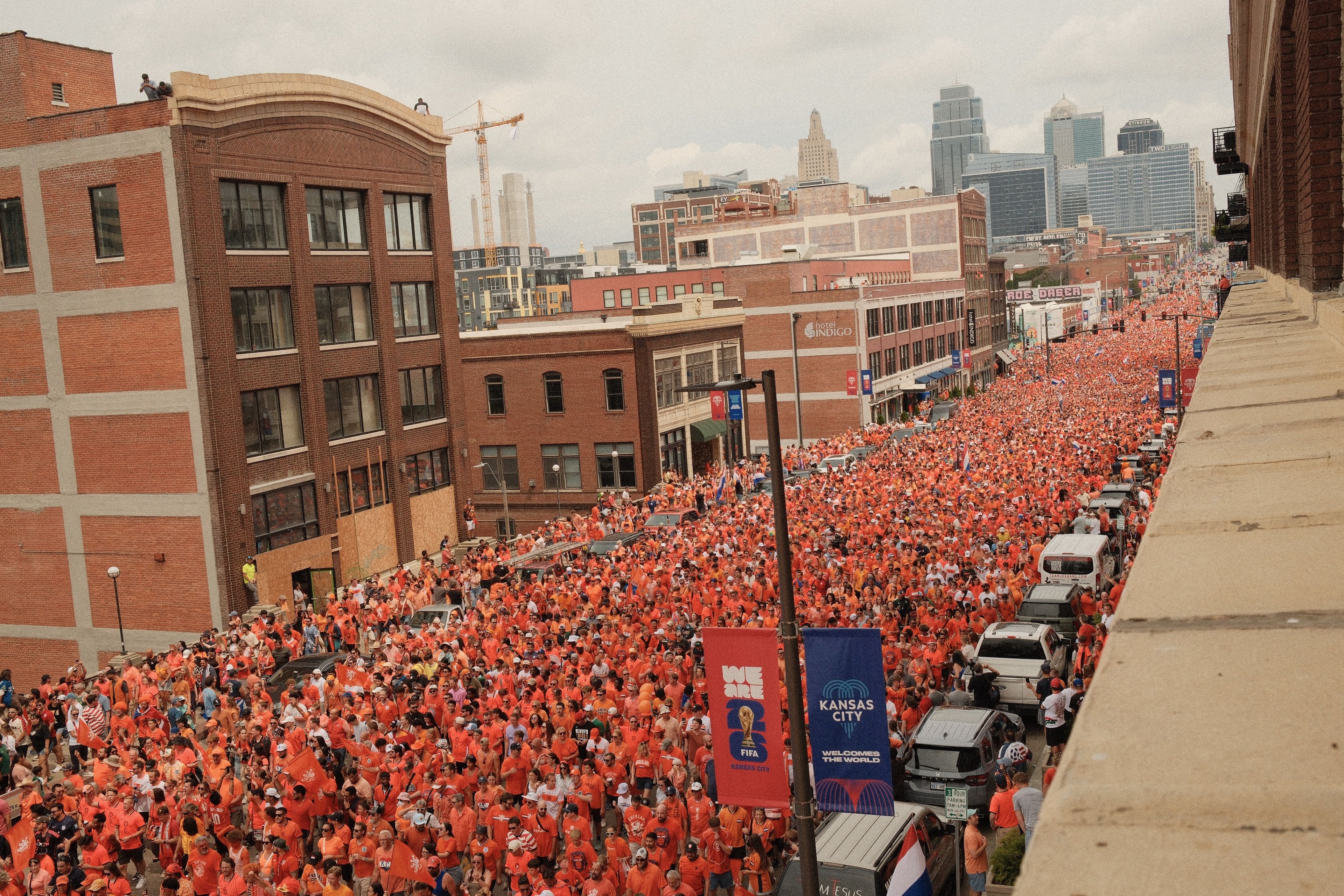 Aerial view of a crowded orange-clad parade on a city street: hundreds of marchers fill the road, brick buildings line the left, Kansas City's downtown skyline in the background, banners and vehicles along the way.