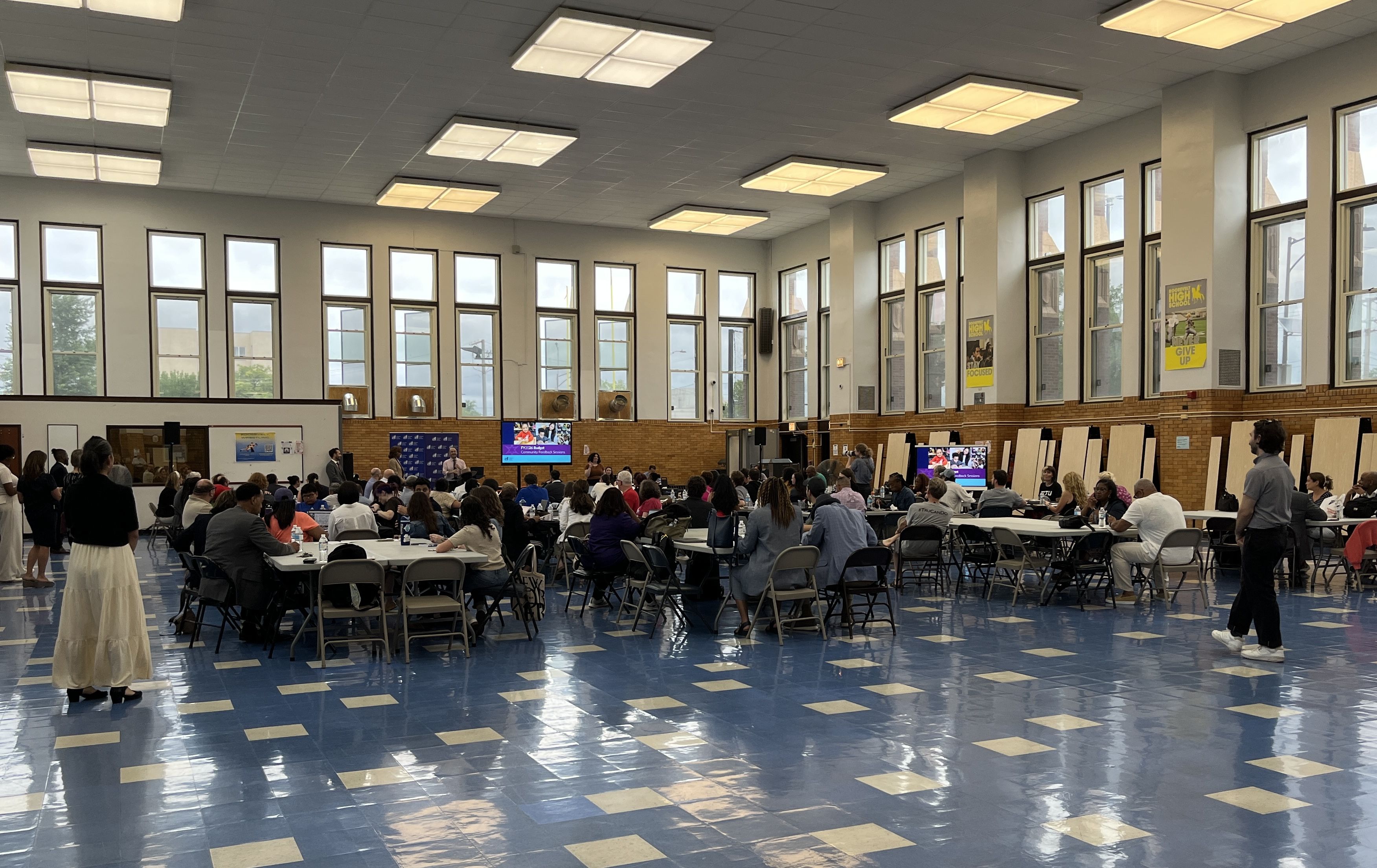 Large group of people seated at round tables in a bright room with tall windows and blue patterned floor, attending a community meeting with screen displays in the background.