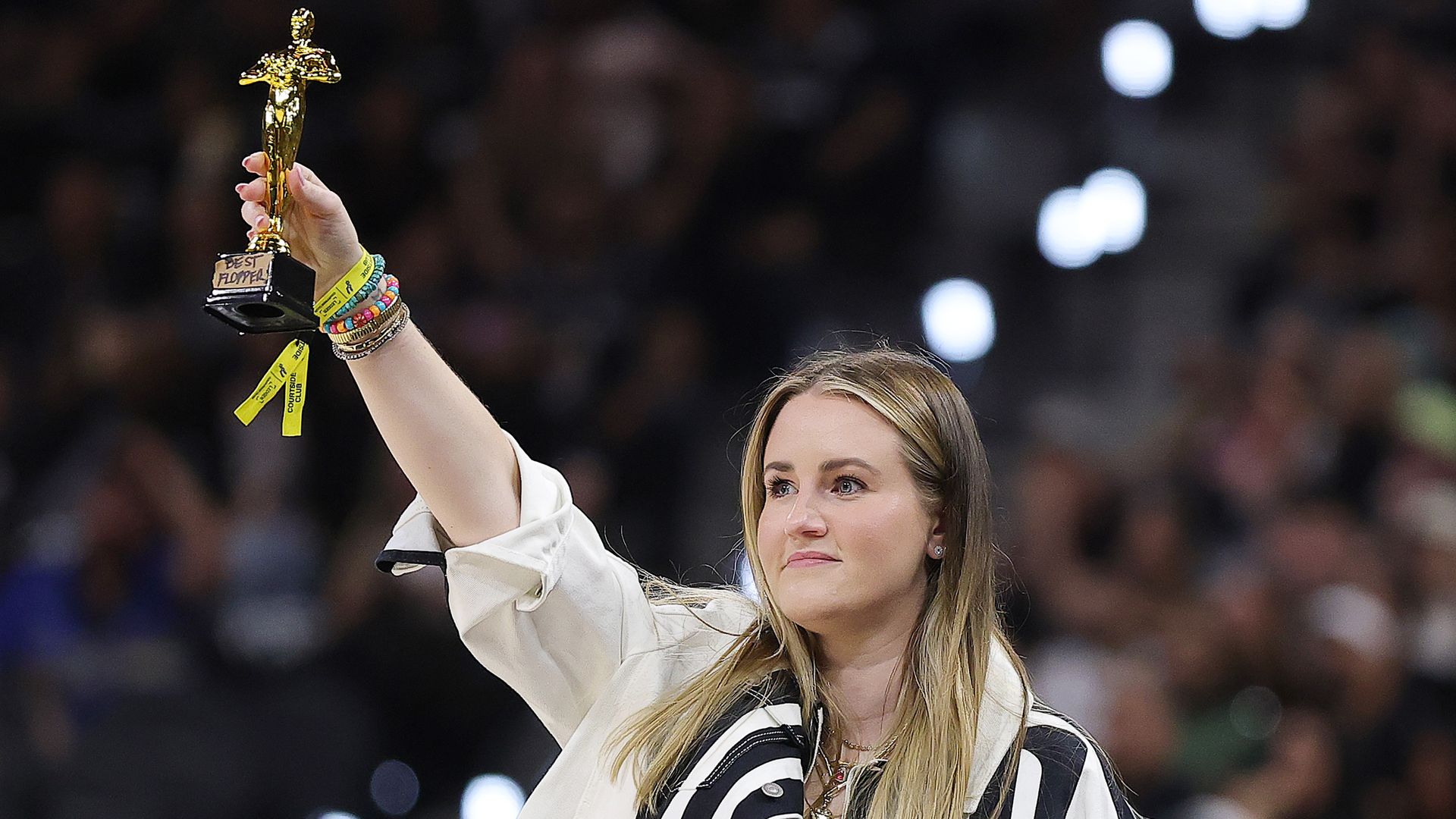 A woman with blond hair raises a gold Oscar statue above her head, wearing a striped black-and-white jacket and colorful bracelets, with a blurred crowd in the background.