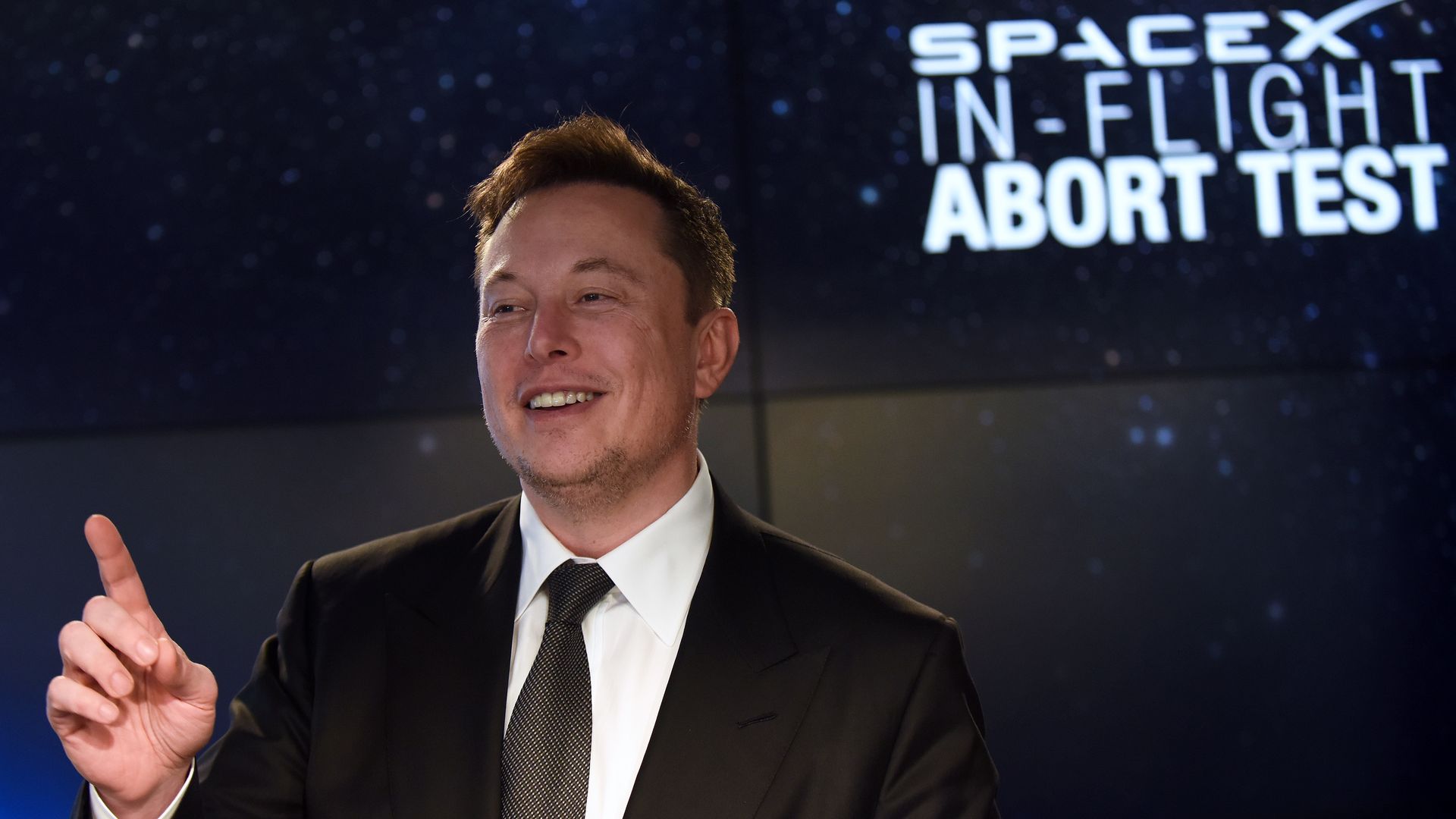 In this image, Elon Musk stands in a suit and tie in front of a large monitor that says "SpaceX in-flight abort test"