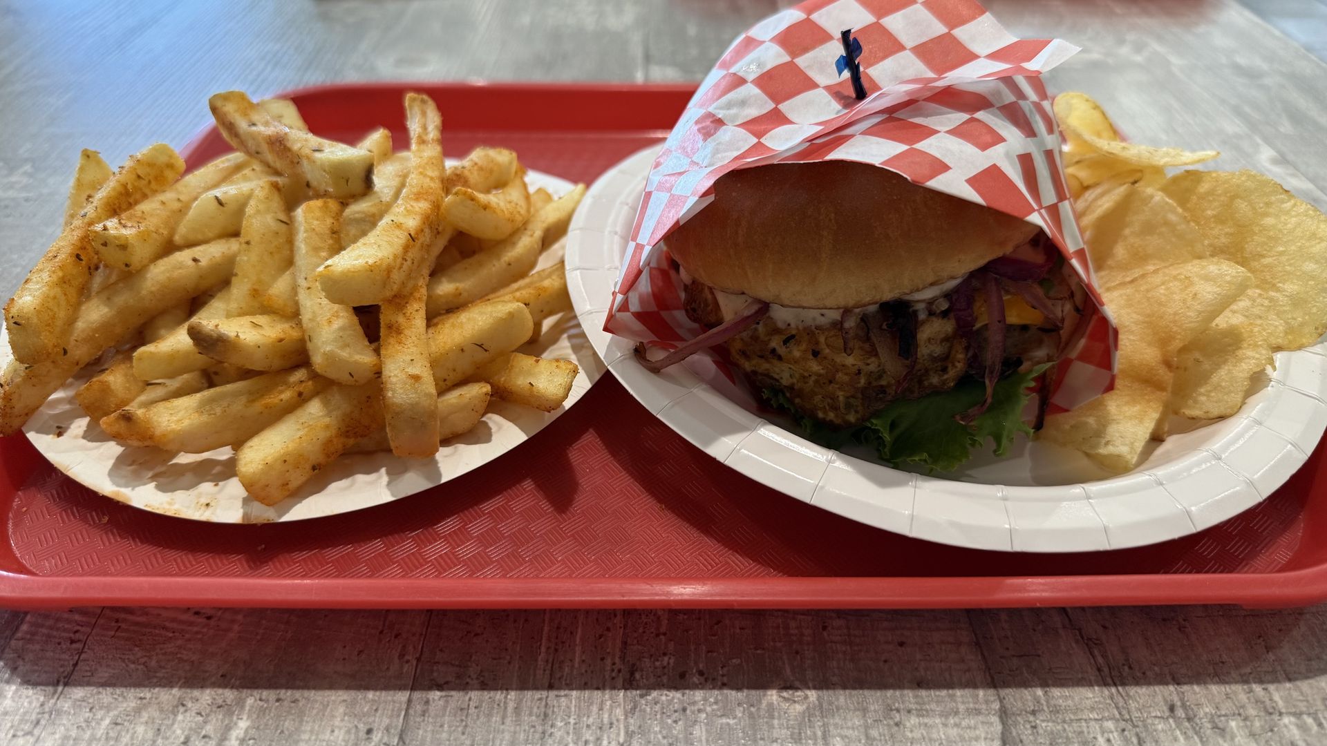 Red tray with a sandwich wrapped partially in red and white checkered paper on a white plate, seasoned French fries on a white plate, and potato chips on the side top right.
