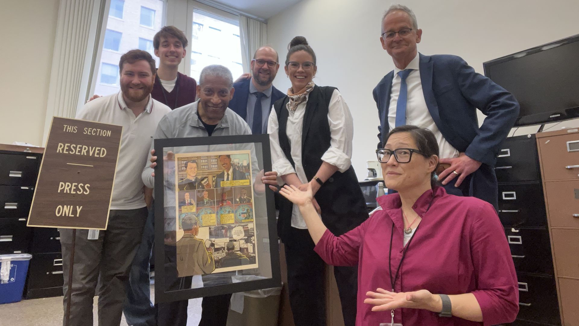 Group of seven people posing in an office, one holding a framed comic-style artwork, another gesturing toward it. A carved wood sign reads "This section reserved press only."