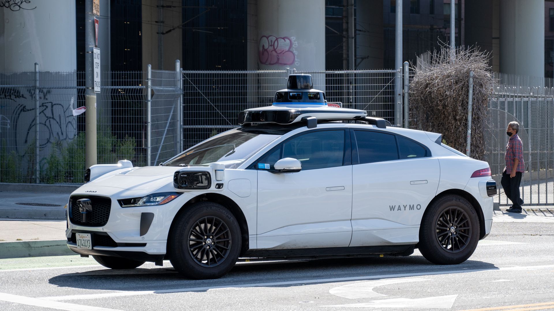 White Waymo self-driving car equipped with sensors on roof and sides, driving on city street with pedestrian nearby under overpass and fenced area in background.
