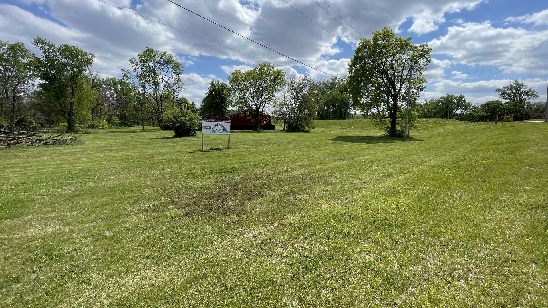 Open grassy park with scattered trees under a partly cloudy blue sky. Overhead power lines cross the scene. A small sign stands in the foreground; a red barn sits among trees in the background.