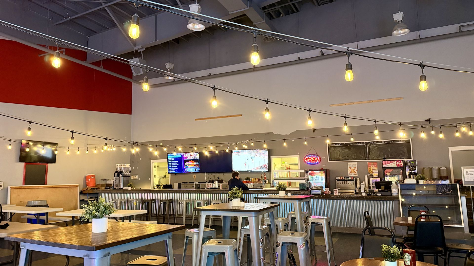 Industrial-style cafe interior with hanging string lights across the ceiling, a red wall on the left, wooden tables and metal stools, a long service counter, and TVs showing a sports game.