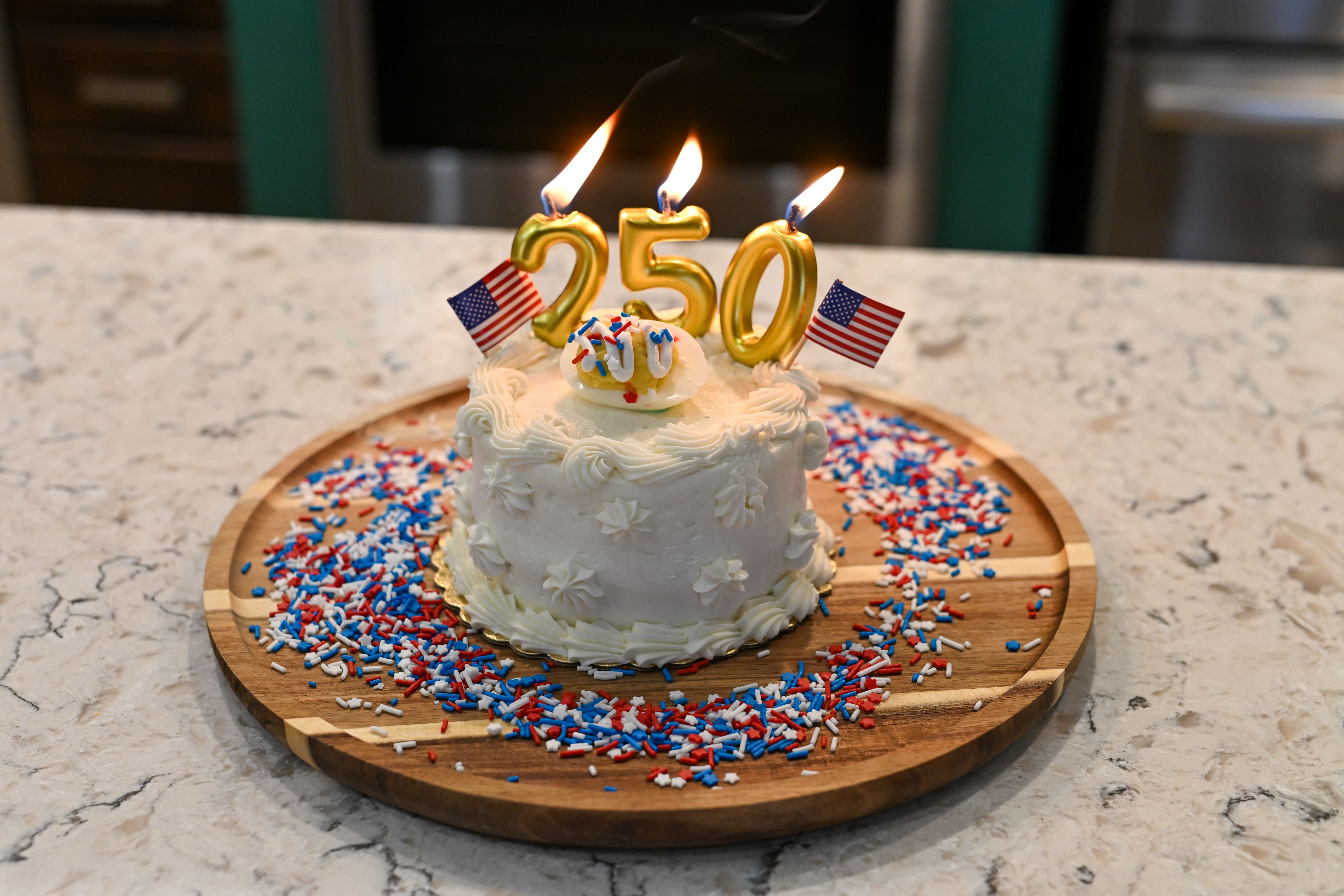 White frosted cake with gold candle numbers 250 lit, small American flags, and red, white, and blue sprinkles on a wooden board atop a marble counter; blurred kitchen background.