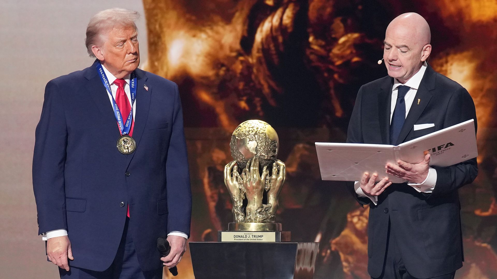  President Trump, wearing a navy jacket with a US flag pin at the top of his left lapel, white shirt, red tie and gold medal, receives the golden FIFA Peace Prize from FIFA President Gianni Infantino, a bald, white man with a black suit, white shirt and black tie, as he reads from a white book. 