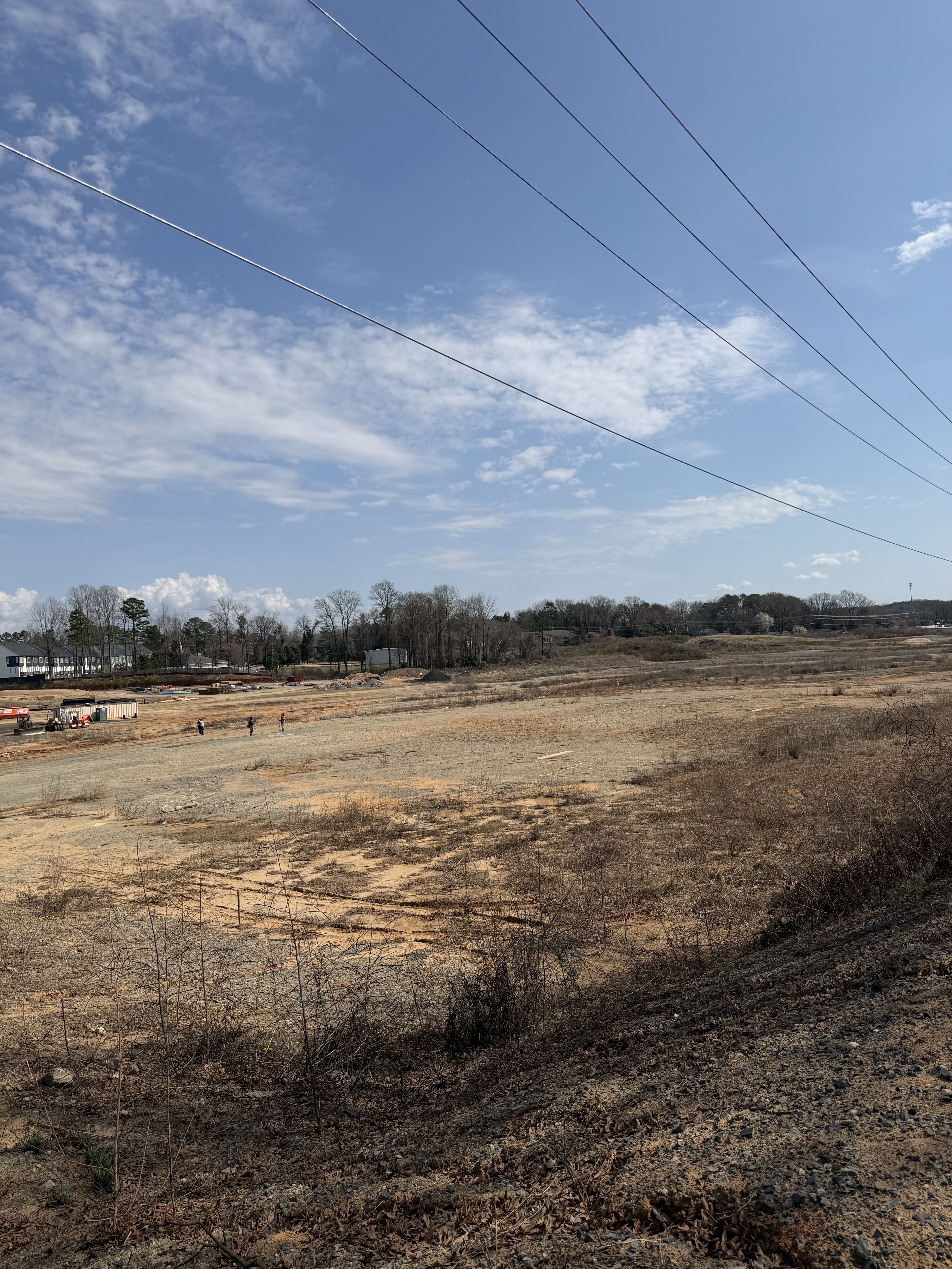 Large clearing with dry brown grass and dirt under a blue sky with light clouds and power lines. Trees and buildings visible in the background with a few people near orange barriers.