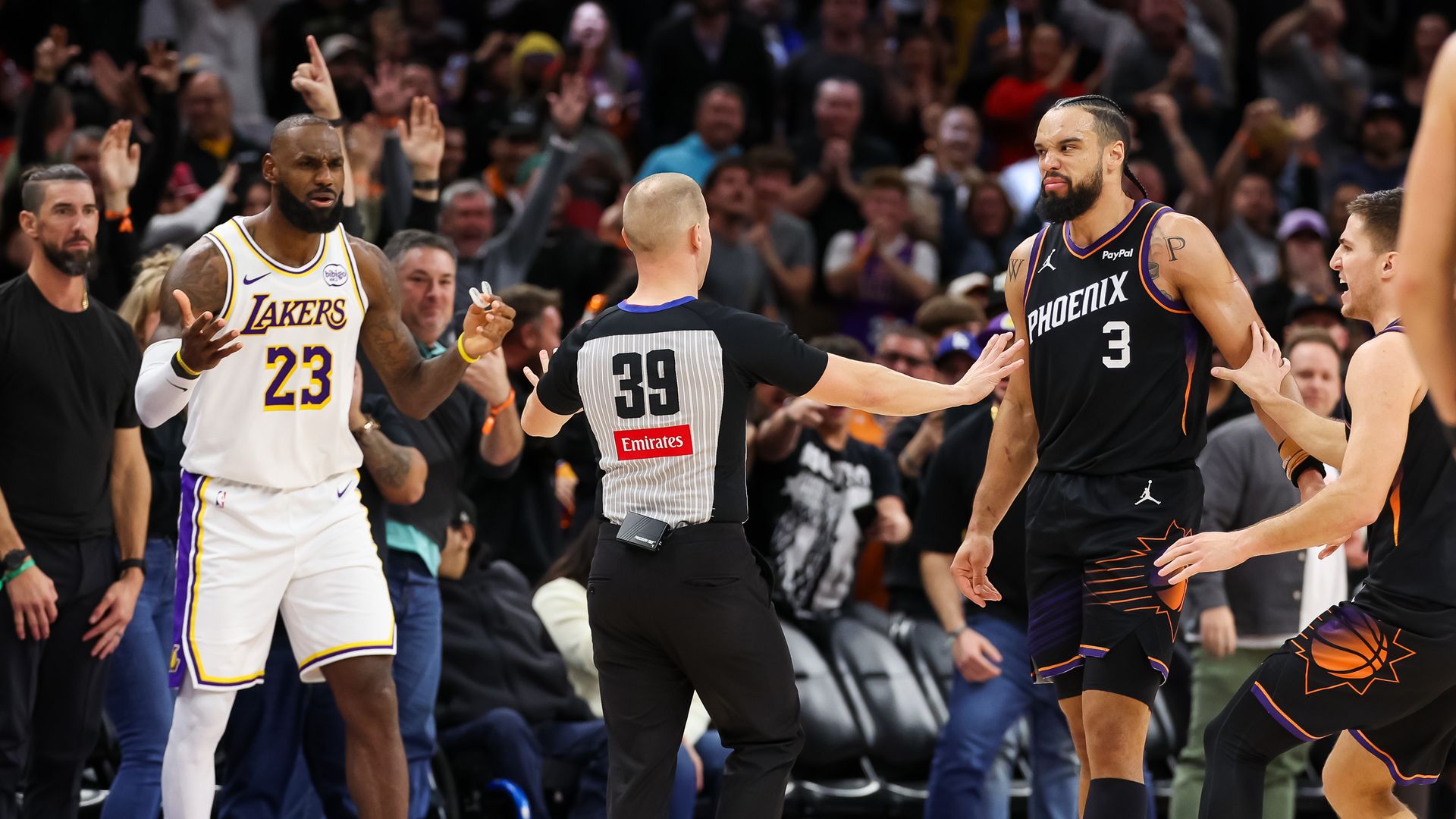 A referee stands between two basketball players, holding his arms out to keep them separate; one player wears a black uniform with "PHOENIX" and the number 3 on the front; the other wears a white uniform with "LAKERS" and the number 23 on the front, holding his hands out in a questioning gesture.