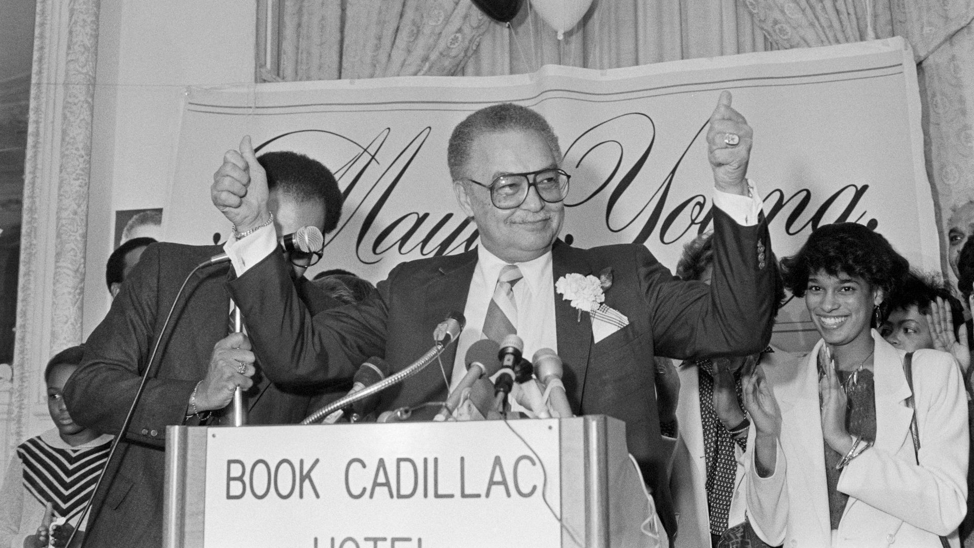 Coleman A. Young stands at a podium with his hands raised, a smile on his face