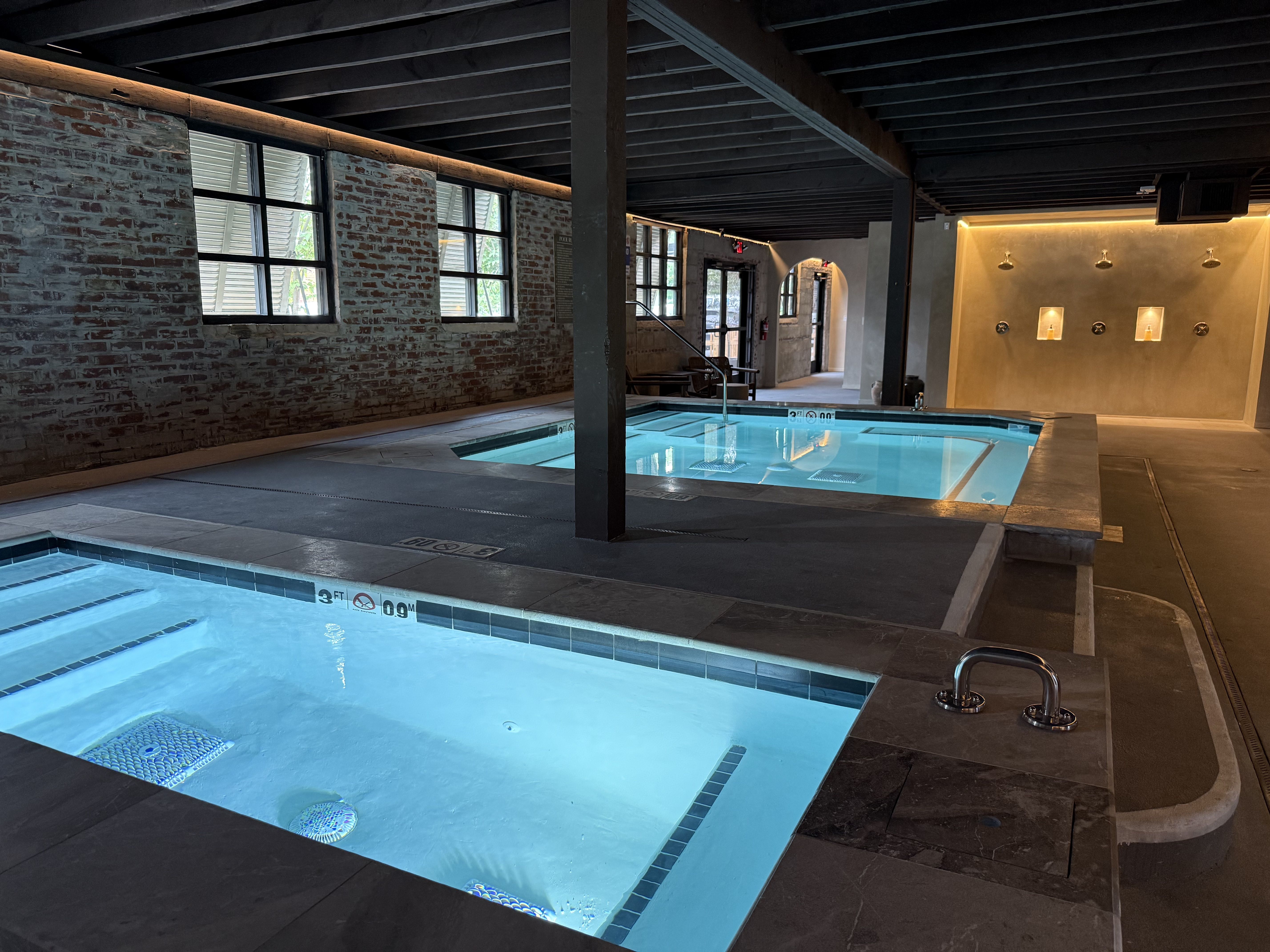 Indoor spa area with two illuminated rectangular pools, exposed brick walls, windows, and warm wall lights on the right side under a dark wooden ceiling.