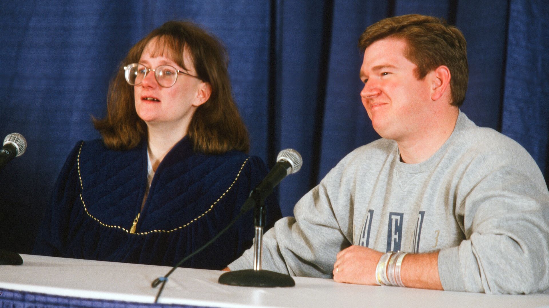 Two people sitting at a table with microphones in front of a dark blue curtain backdrop. Woman with glasses and dark blue jacket; man in a light gray sweatshirt with wristbands.