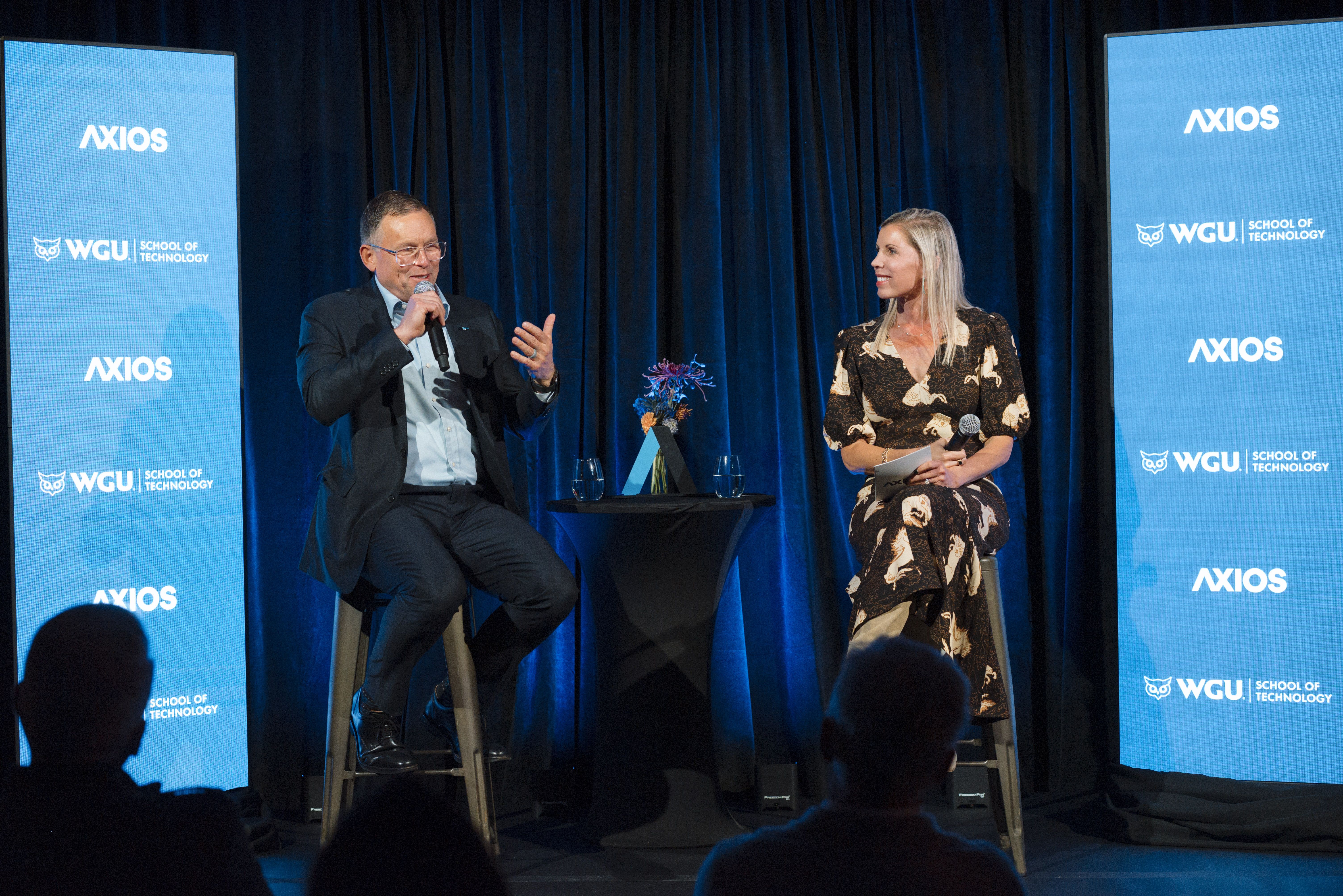 Two panelists on stage with blue Axios screens and WGU logos; man in a suit speaks into a handheld mic, woman in a floral dress holds a mic and notebook; audience silhouettes in foreground.