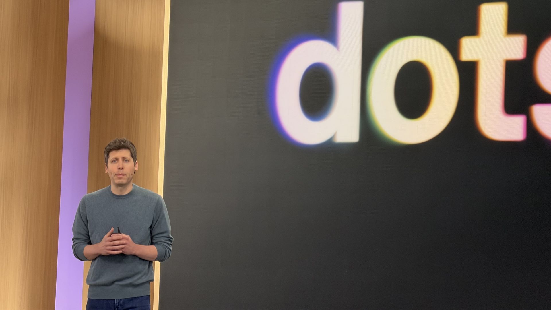 Sam Altman wearing a gray sweater stands on a stage in front of a large black screen with oversized rainbow-edged letters; wooden wall panels and a purple vertical stripe are on the left.
