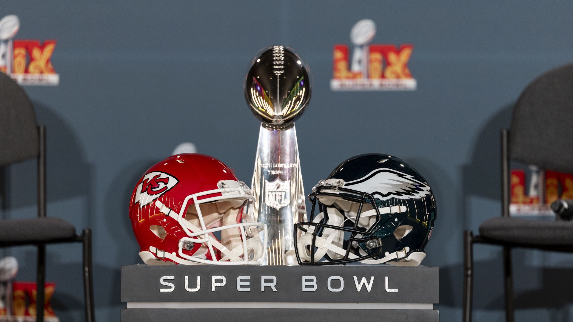 Two American football helmets sit side by side while divided by a shiny metal trophy. The word SUPER BOWL is written on the bottom.