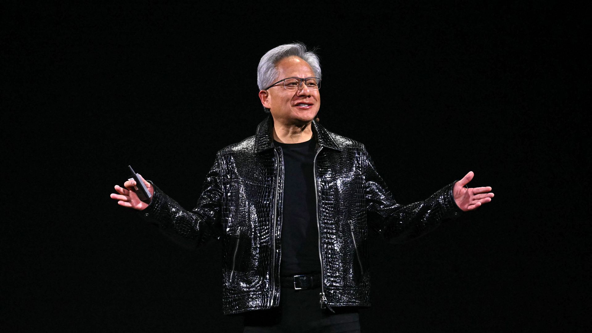 A man in a black leather jacket gestures with his arms apart, standing in front of a black background.