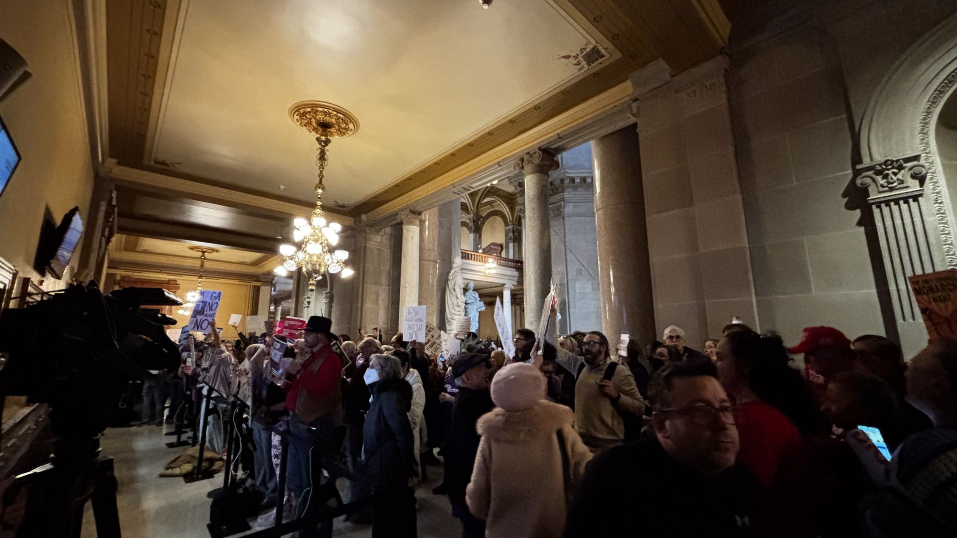 Crowd of diverse people gathered inside a grand building with tall marble pillars, ornate ceiling, and chandeliers, holding protest signs and recording with phones in dim lighting.