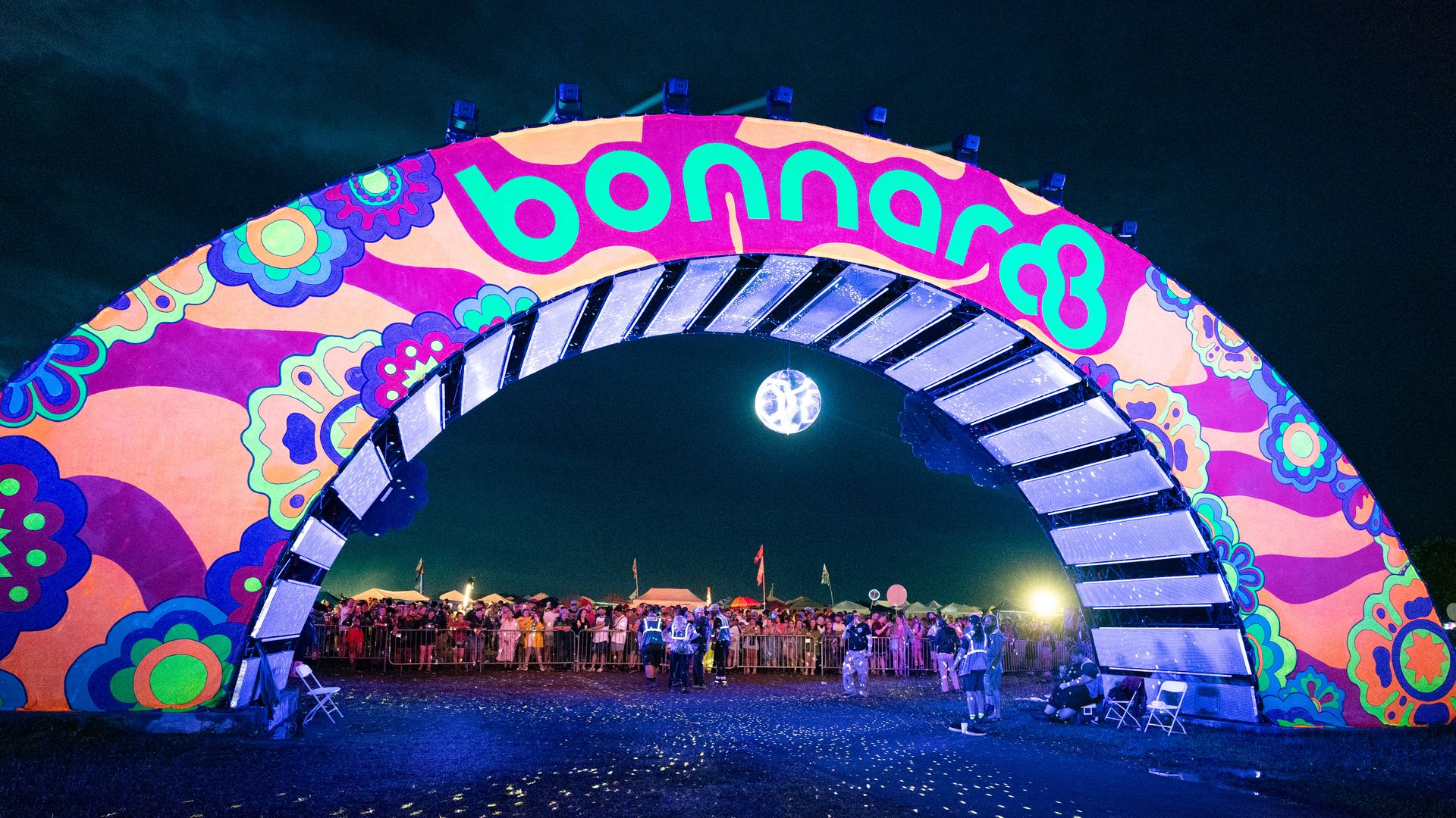 Fans stand under a lit "Bonnaroo" arch filled with cartoon flowers