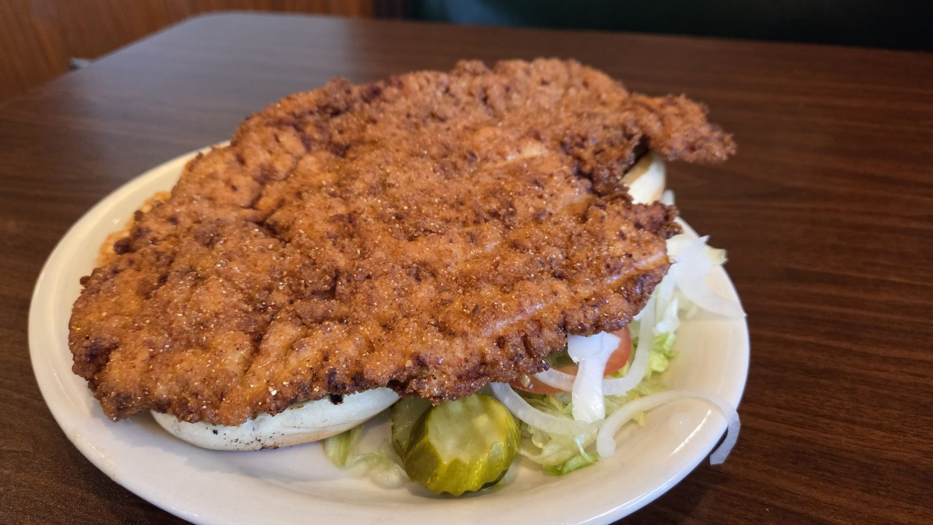 Plate with a large fried chicken sandwich featuring crispy brown breading, sliced pickles, onions, lettuce, and a toasted bun, on a wooden table background.