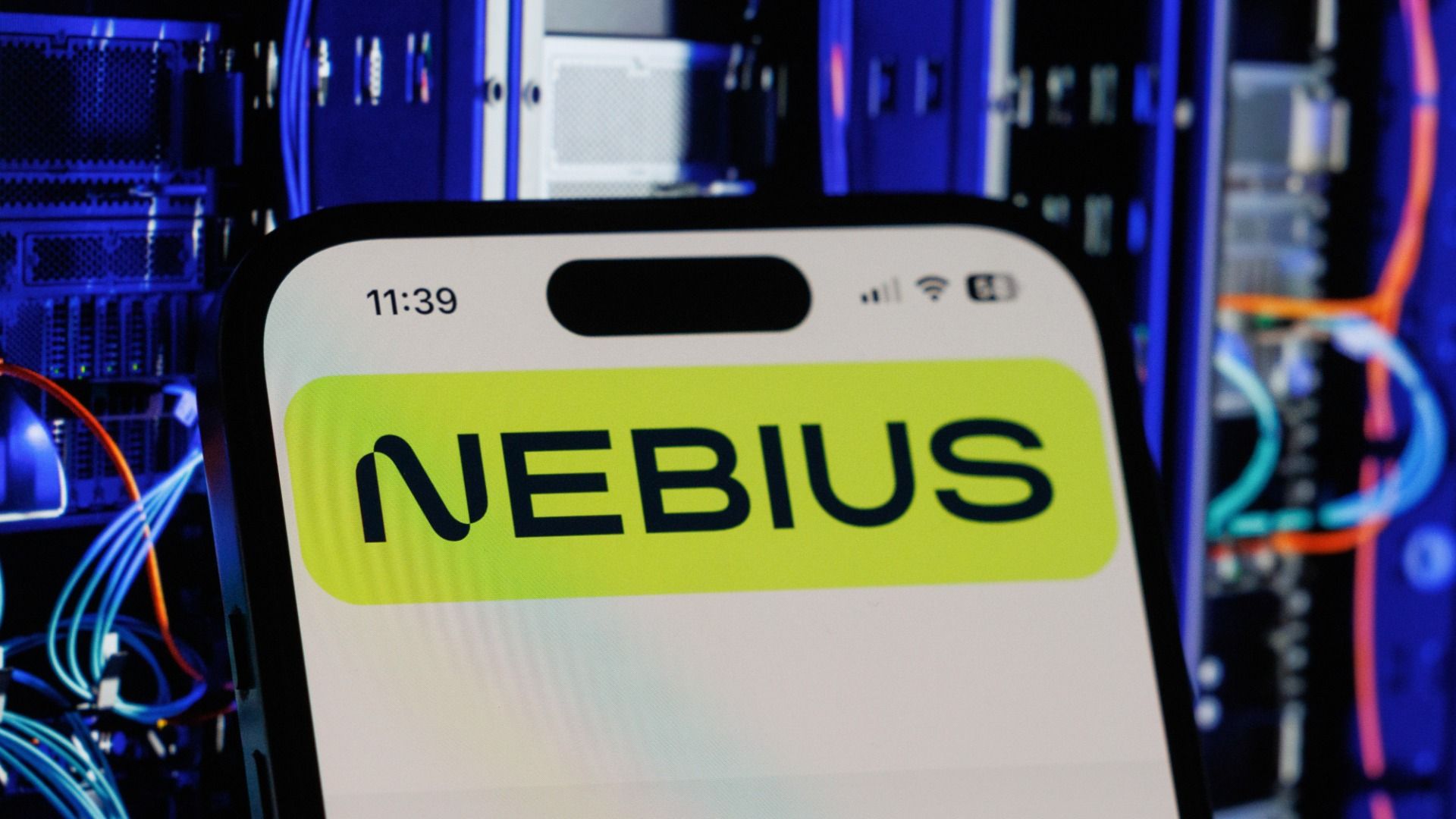 A Nebius Group NV logo on a smartphone arranged in front of a blurry background of blue and red servers and wires in Riga, Latvia.