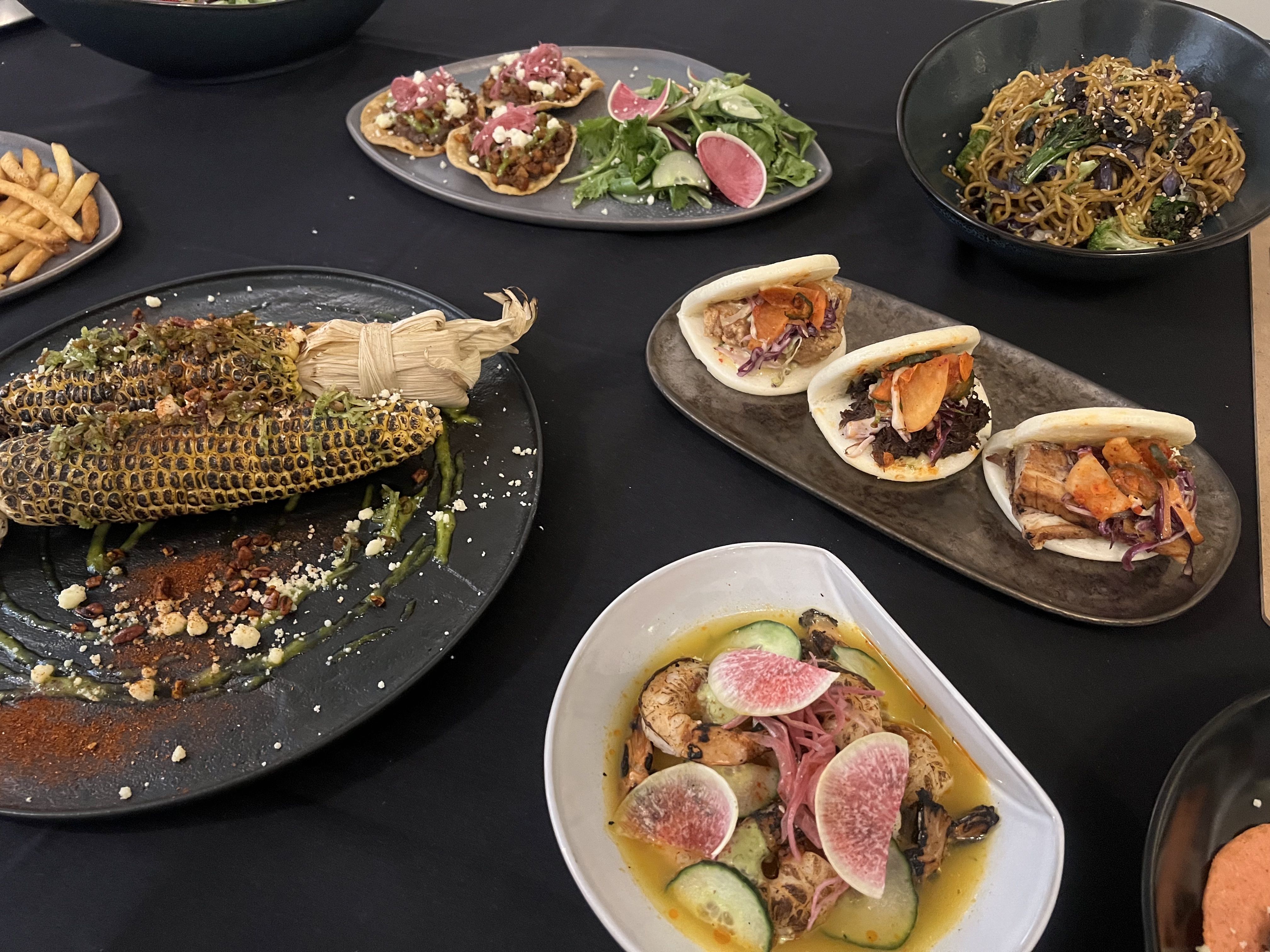 Various dishes on a black table including grilled corn with seasoning, three bao buns with fillings, a salad with radishes and cucumbers, small tostadas, a bowl of noodles with vegetables, and fries.