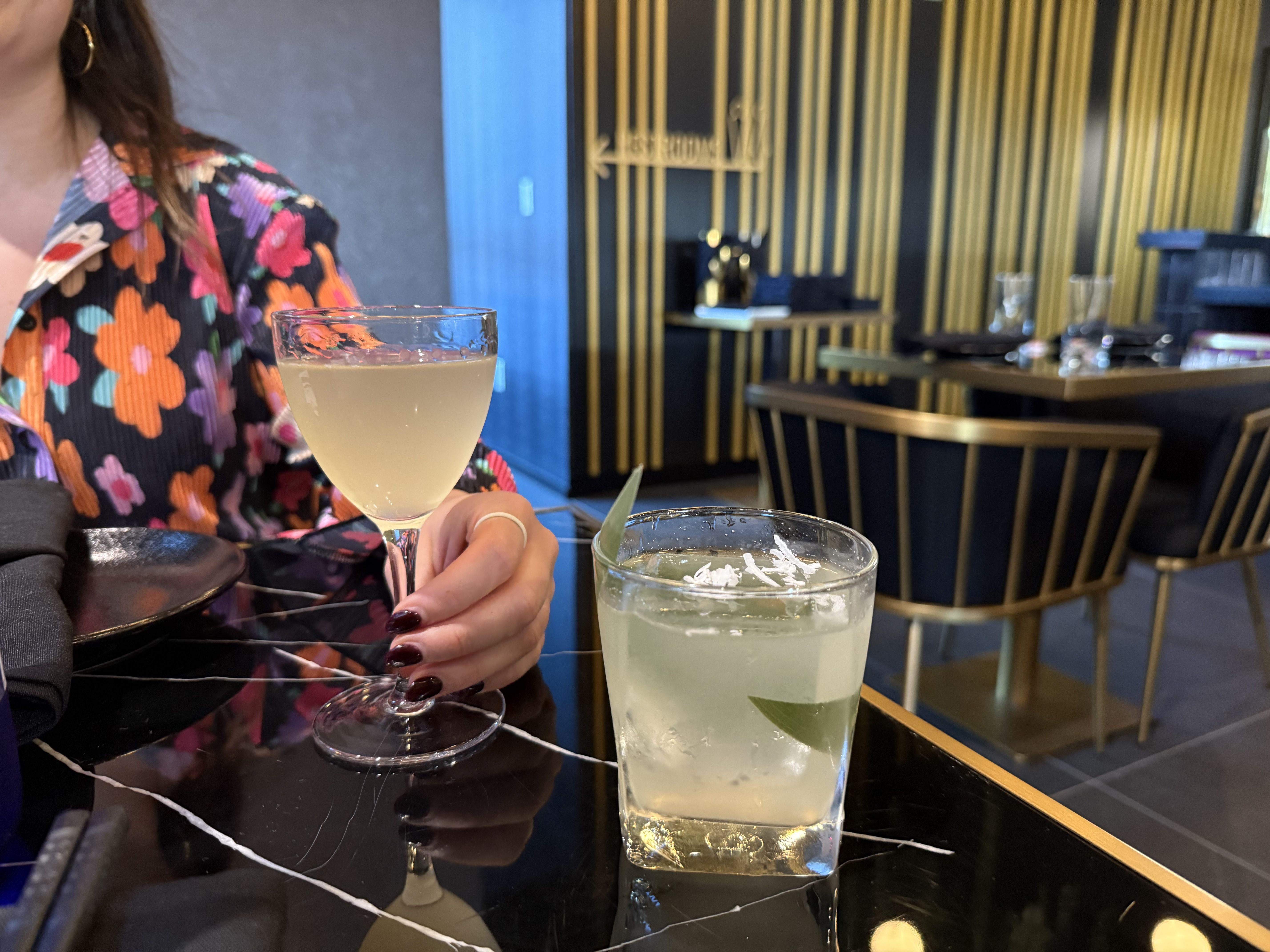 Person with dark floral shirt holding a light-colored cocktail in a stem glass on a black marble table with a short glass cocktail garnished with leaves, in a modern bar with gold-striped walls.