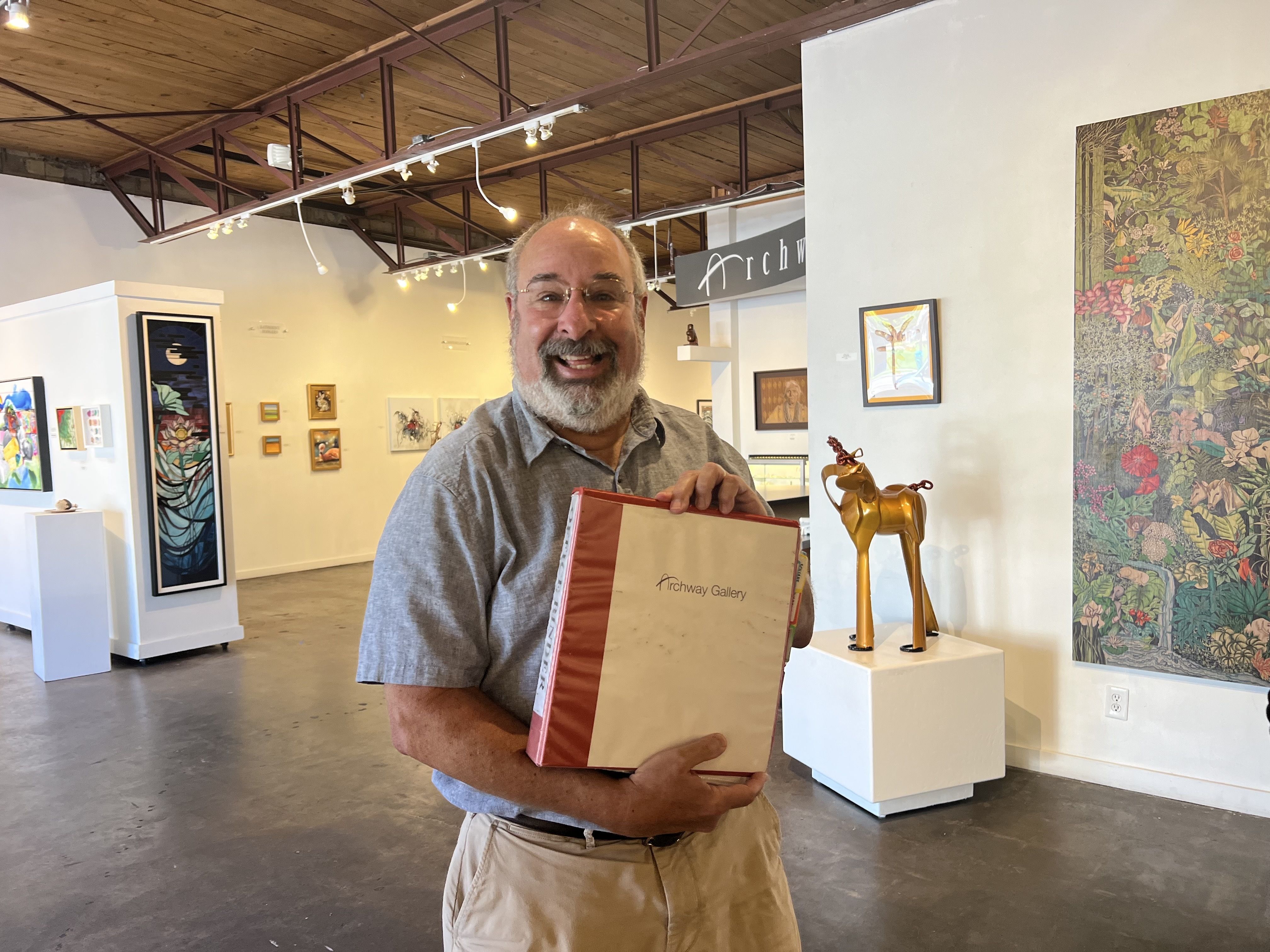 Smiling man with glasses and beard holding an "Archway Gallery" binder inside an art gallery with paintings and sculptures visible.
