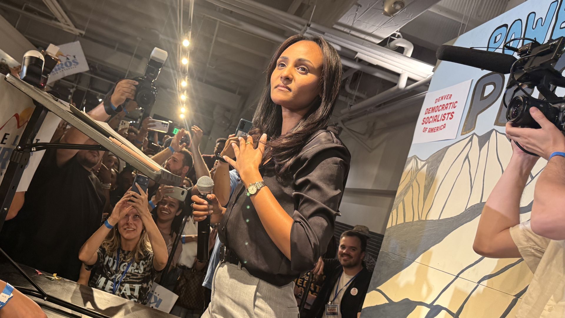 A woman in a blazer speaks at a podium as reporters and photographers surround her; a right-side banner reads "Denver Democratic Socialists of America".