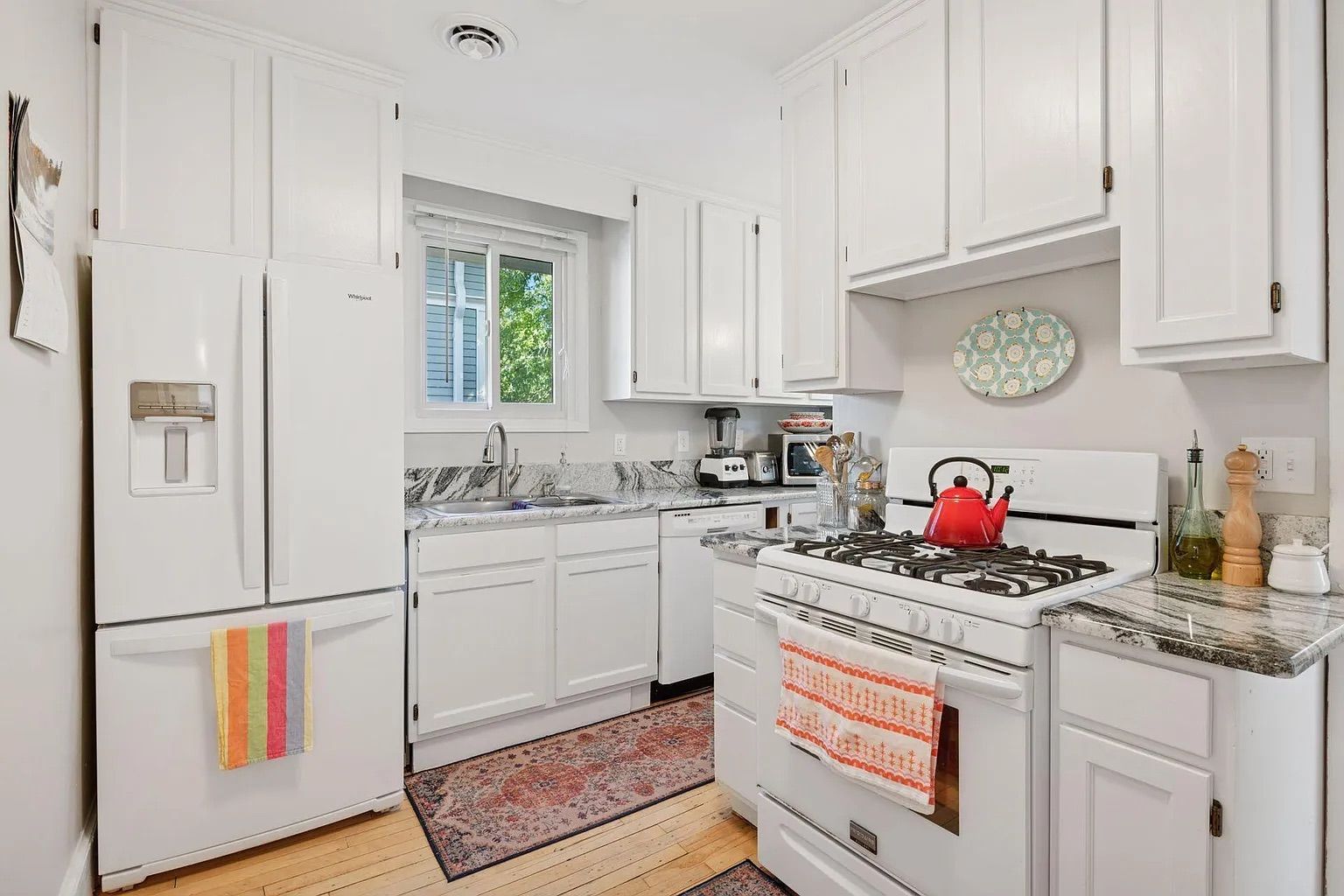 Bright white kitchen with marble countertops, white cabinets, a white Whirlpool fridge with a colorful striped towel, a white gas stove with an orange towel and red kettle, and patterned rugs on wood floor.