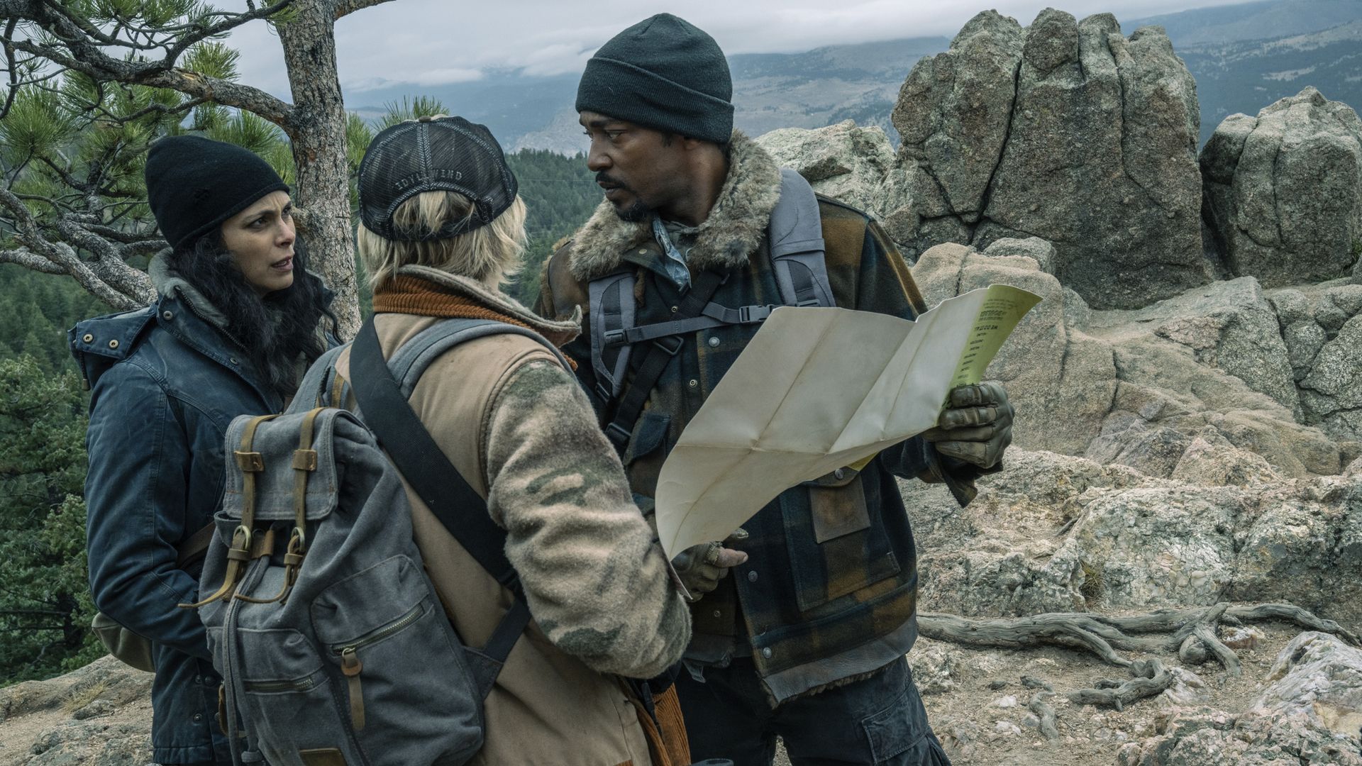 A man in a beanie and heavy jacket holds up a map while speaking with two women. 
