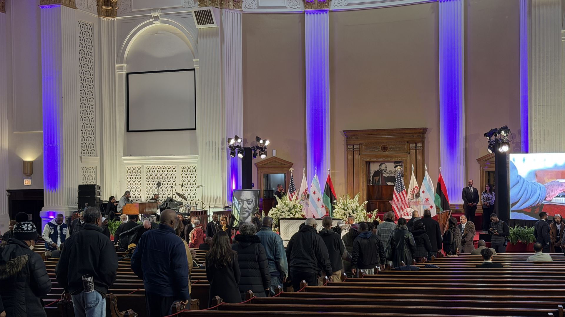 People gathered in a large hall with tall columns lit purple, standing in line near a white casket surrounded by white flowers and flags, including U.S. and Chicago flags, and a portrait.