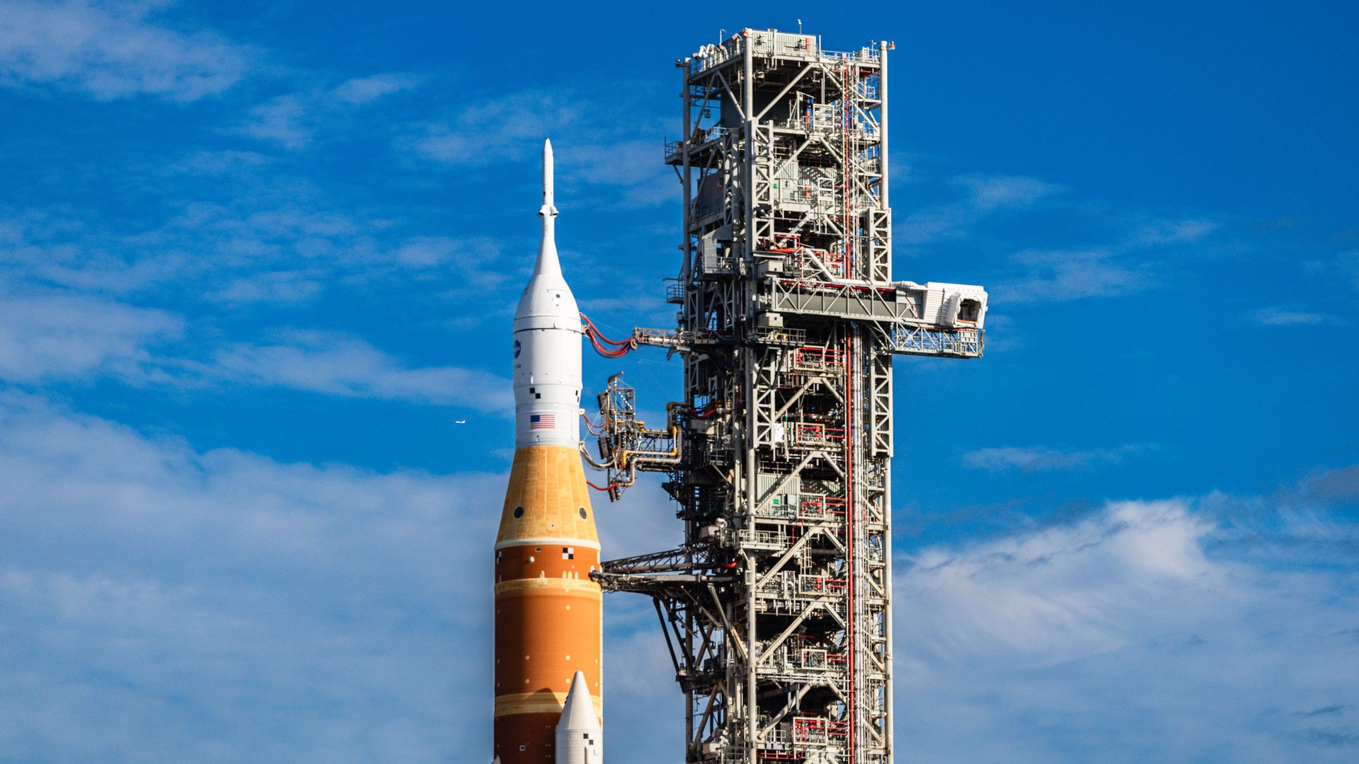 Orange and white NASA rocket on launch pad with tall metal scaffold against blue partly cloudy sky.