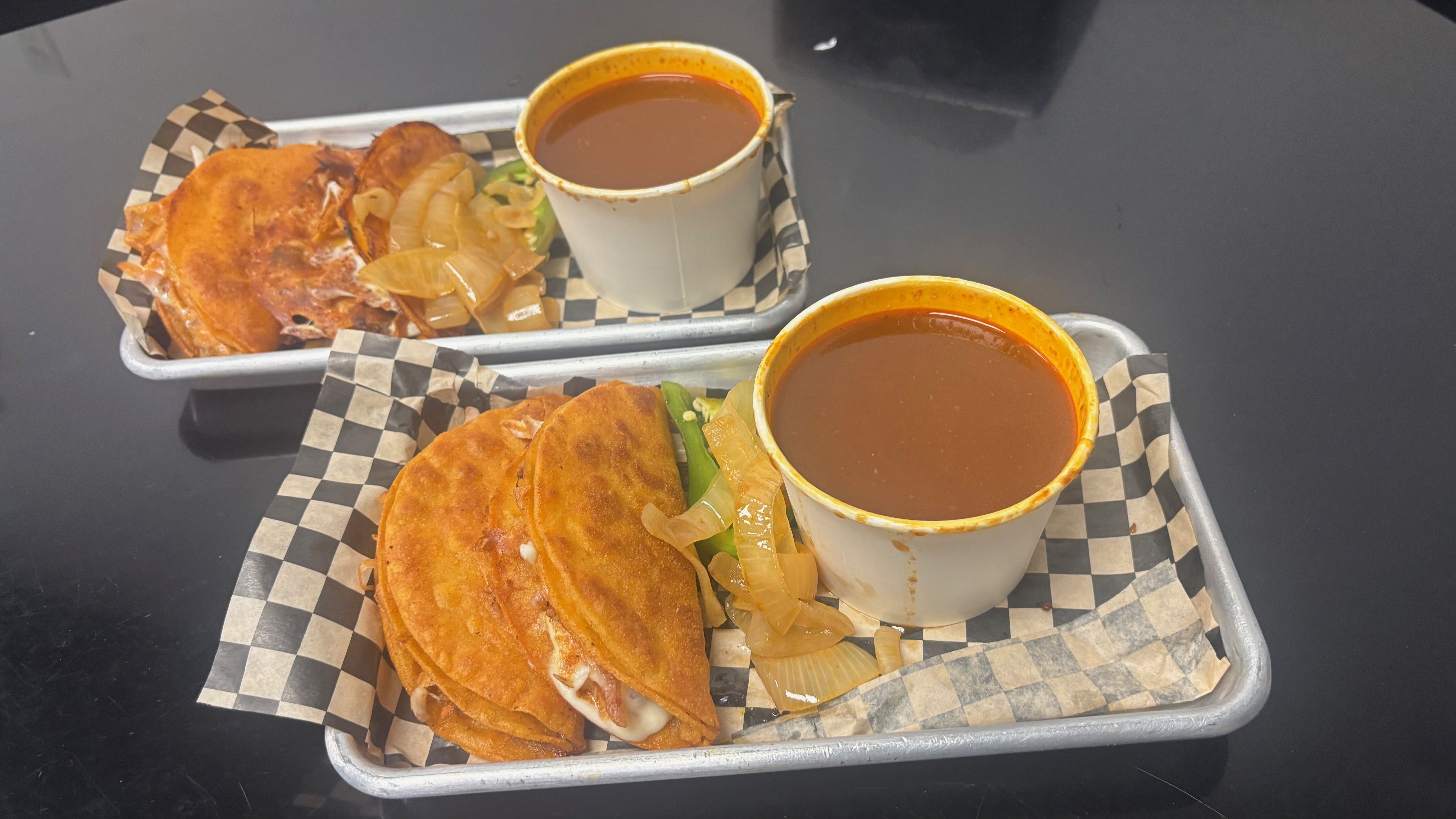 Two trays with three golden-brown fried tacos each, topped with sautéed onions and jalapeños, accompanied by cups of red dipping sauce on checkered paper, set on a black table.