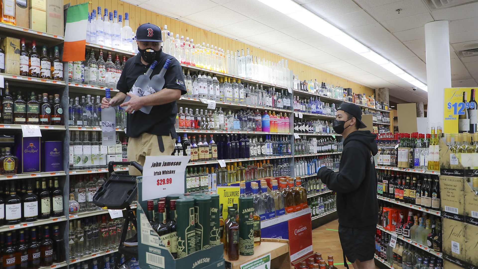 A man wearing shorts and a face mask stands near another man in a face mask, who is standing on a ladder in front of shelves of alcohol 