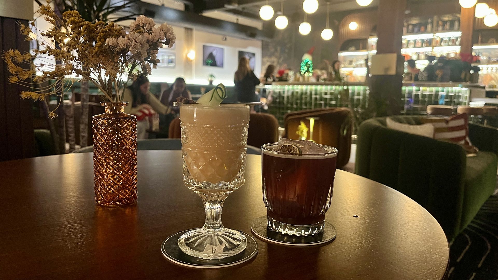 Two cocktails on a wooden table in a dim bar with green and brown chairs, a brown vase with dried flowers, and people at the illuminated bar in the background.
