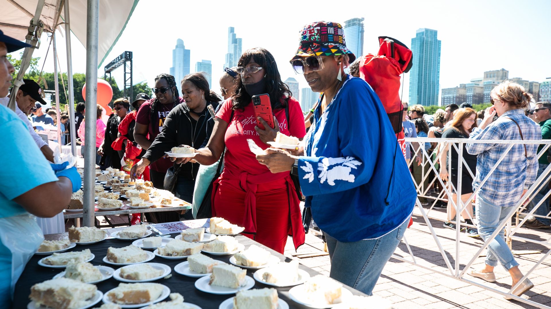 A line of people stand in front of a table with slices of cheesecake on paper plates with the Chicago skyline in the background. 
