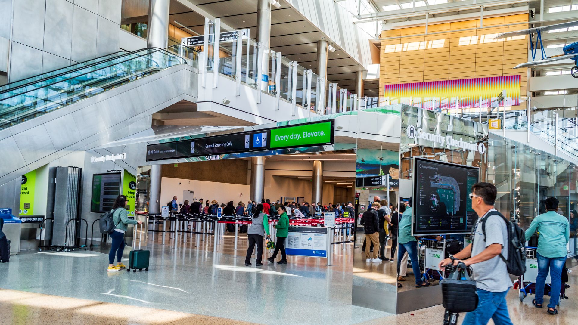 A view of a security checkpoint with people entering the lines preceding the checkpoint and people with luggage walking toward the checkpoint, with an escalator at left and an upper level that is open.
