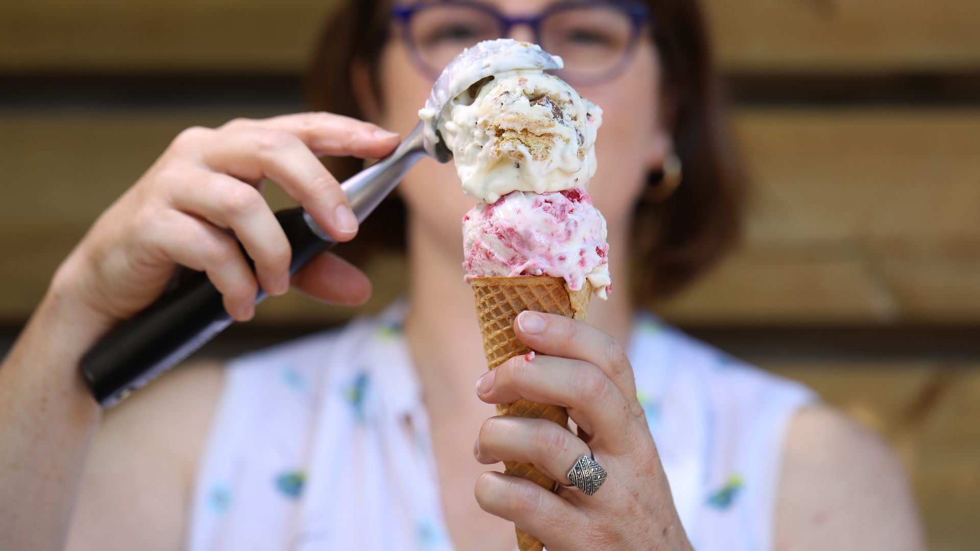 Person with blue glasses holds a waffle cone with two ice cream scoops while a spoon is near the scoop, against a wooden background.
