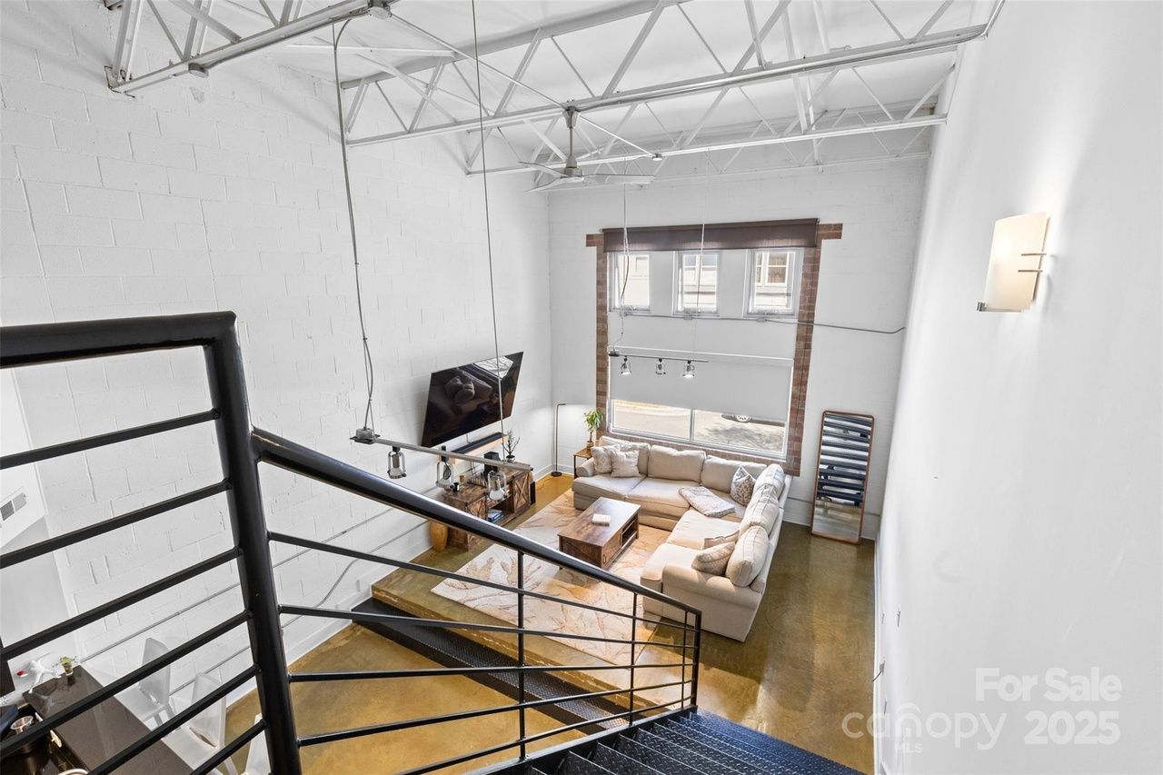 Bright loft living room seen from staircase, featuring white walls, high ceilings with exposed beams, beige sectional sofa, wall-mounted TV, large window, and polished concrete floor.
